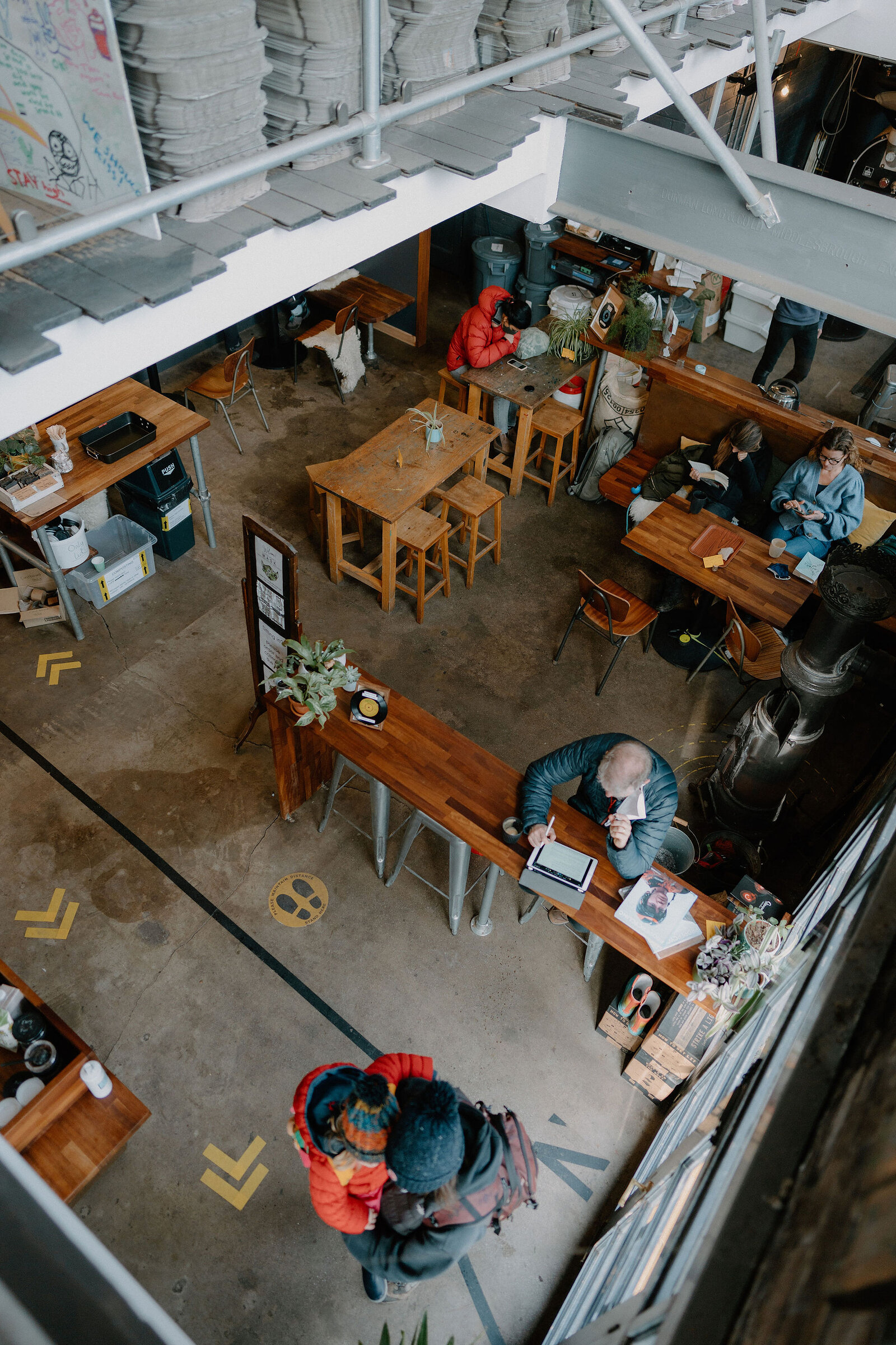 An aerial view looks down over a busy industrial-style cafe, with customers seated at wooden tables and high stools.