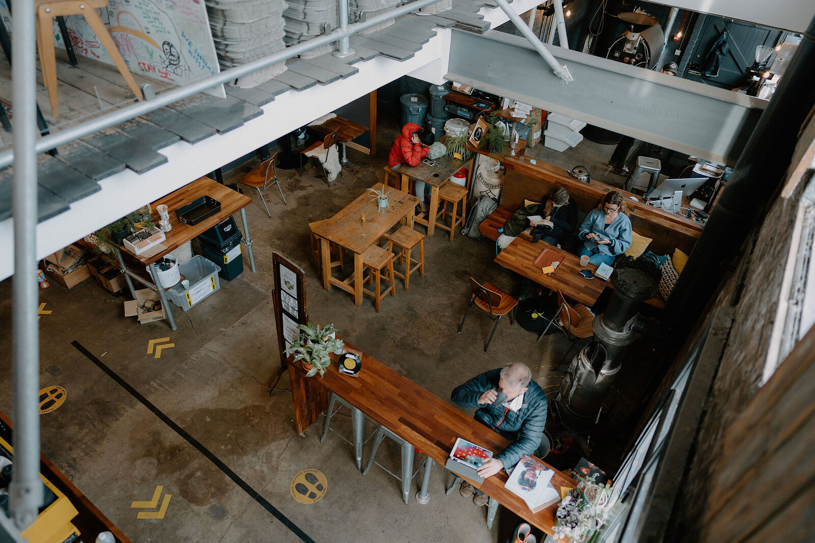 Customers sit at wooden tables in a busy industrial-style cafe, seen from a mezzanine above.