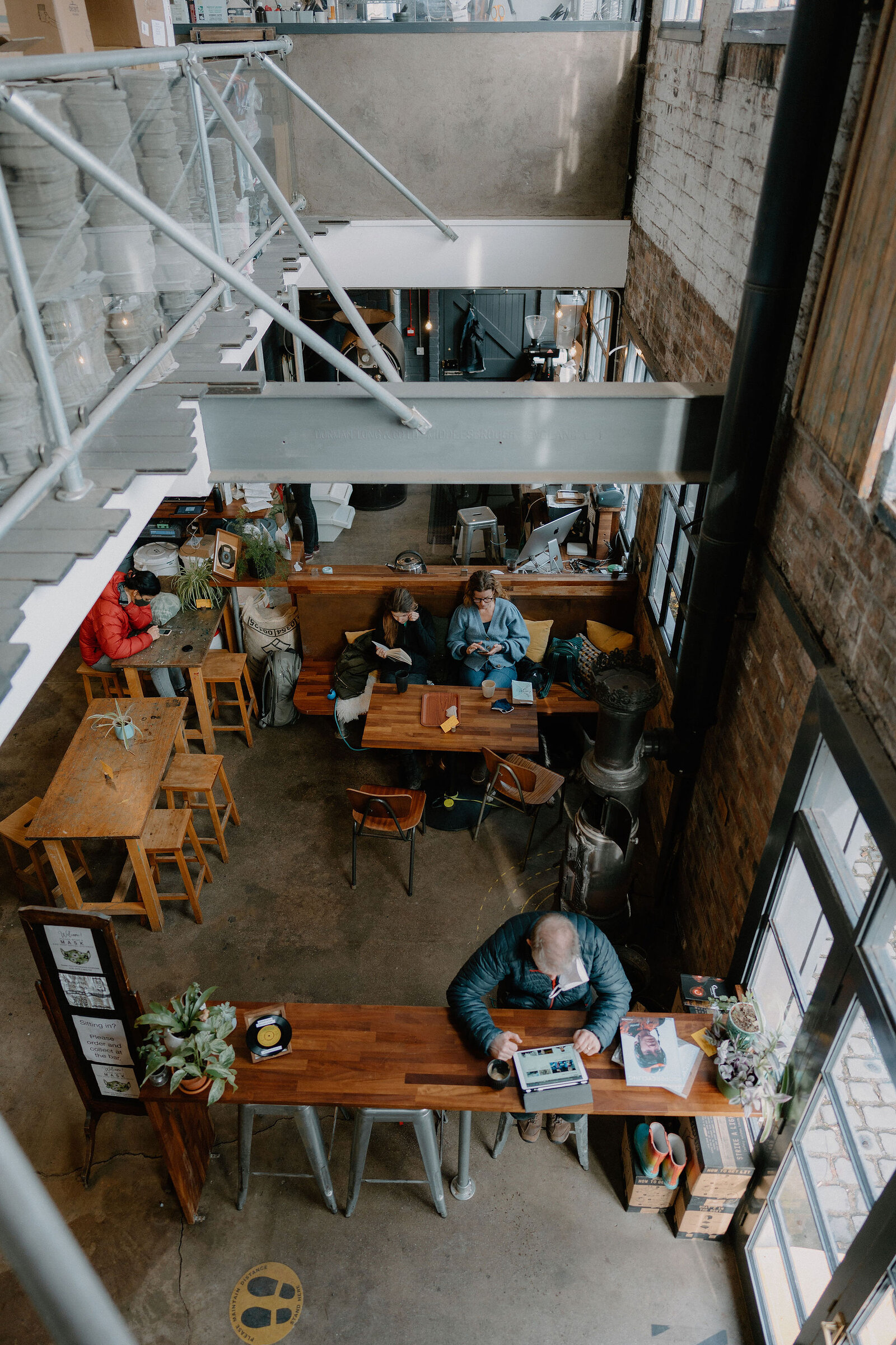 Customers sit at wooden tables in a busy industrial-style cafe beneath exposed pipework and a mezzanine walkway.