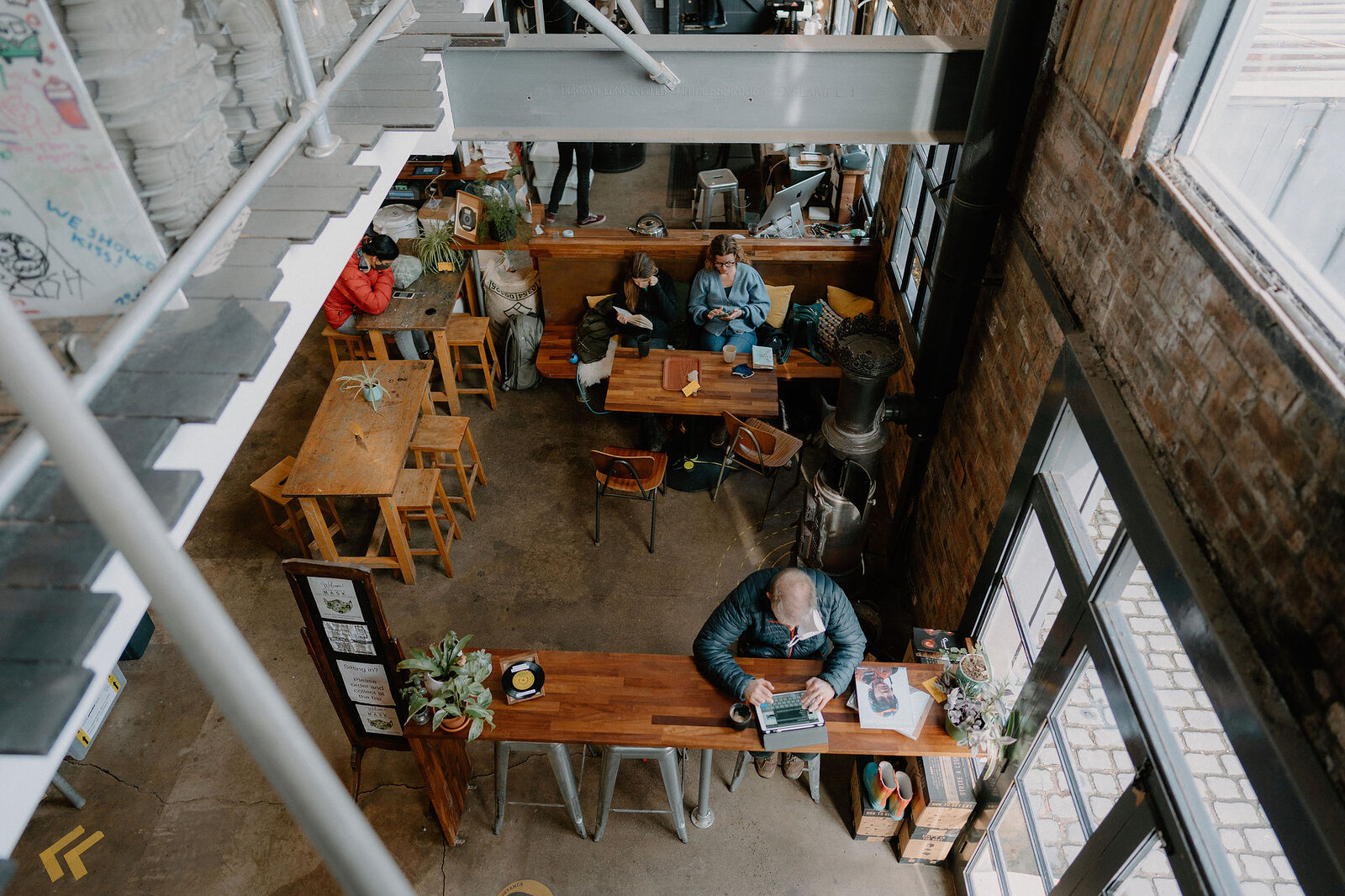 Customers sit at wooden tables in a busy industrial-style cafe, seen from above through a metal-framed mezzanine.