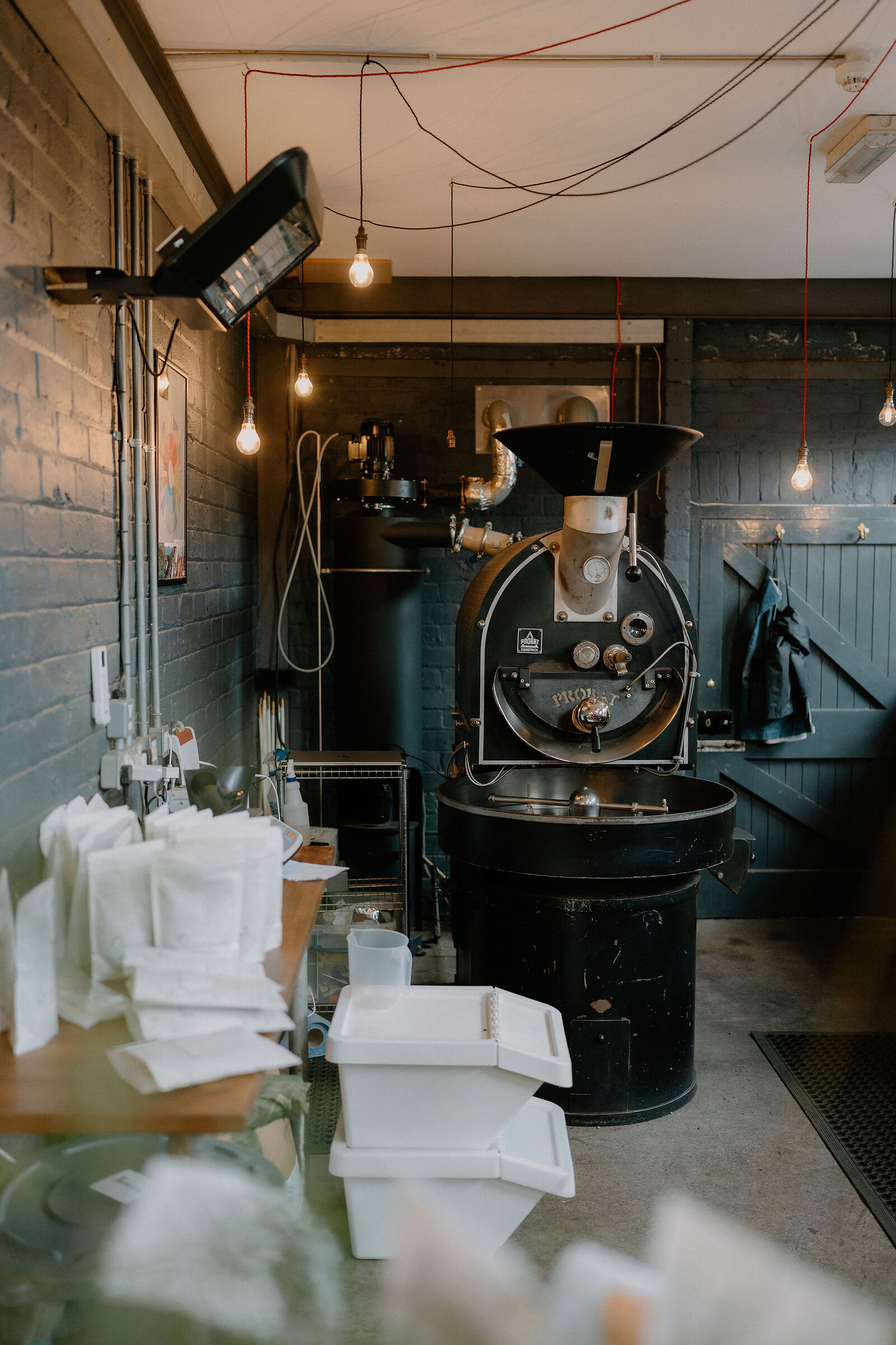 A large coffee roasting machine stands in a dark-walled roastery, bags of beans stacked on the counter beside it.
