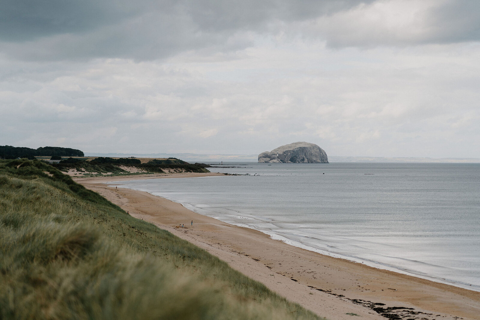 Bass Rock rises from calm water beyond a sandy beach edged with dunes.