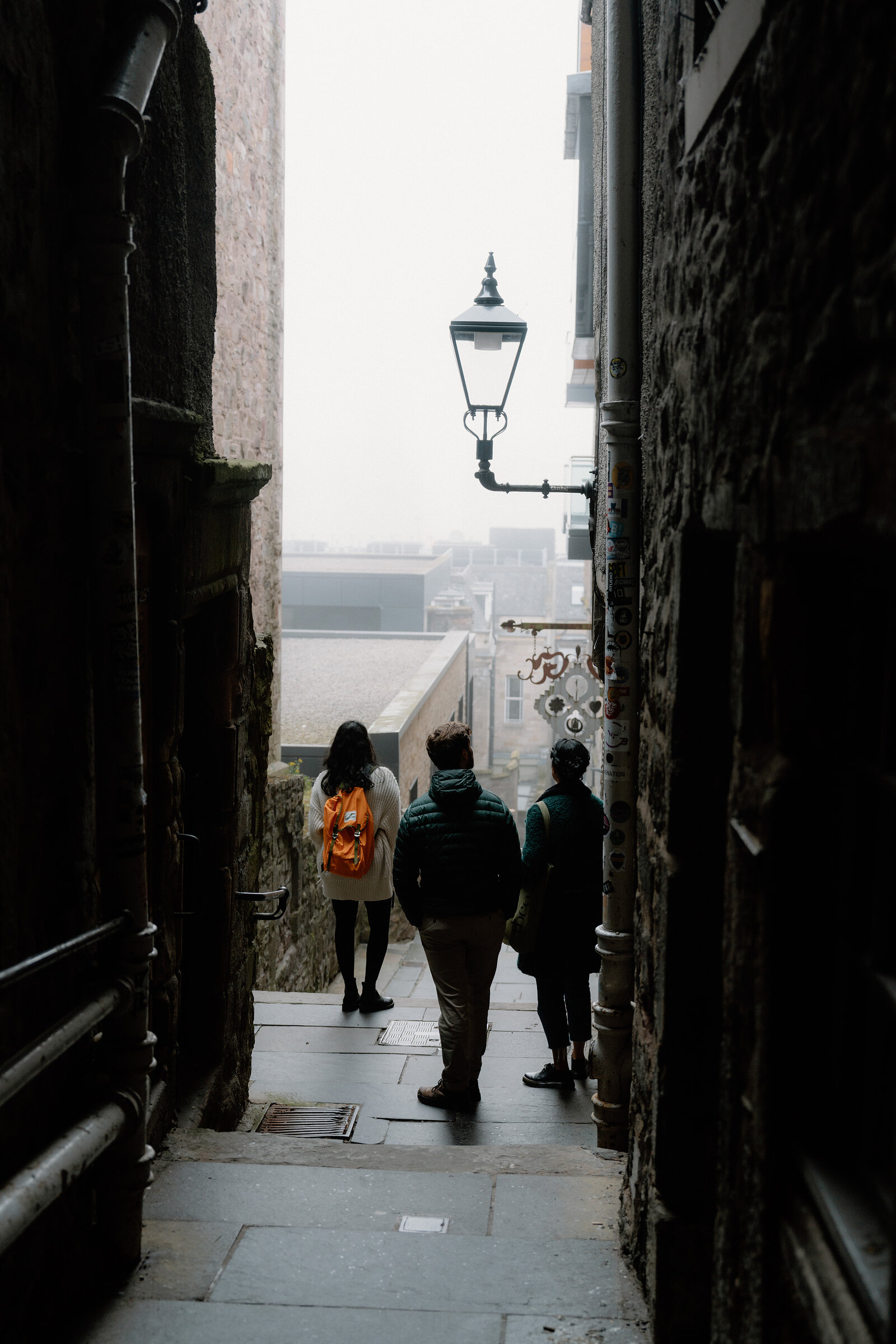 Three people walking up a narrow close towards a hanging lamp and rooftops beyond