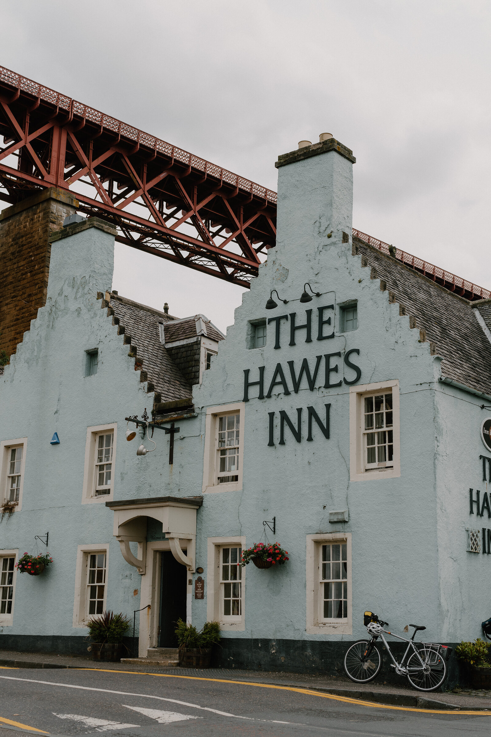 The pale blue gabled facade of The Hawes Inn beneath the Forth Bridge's red girders