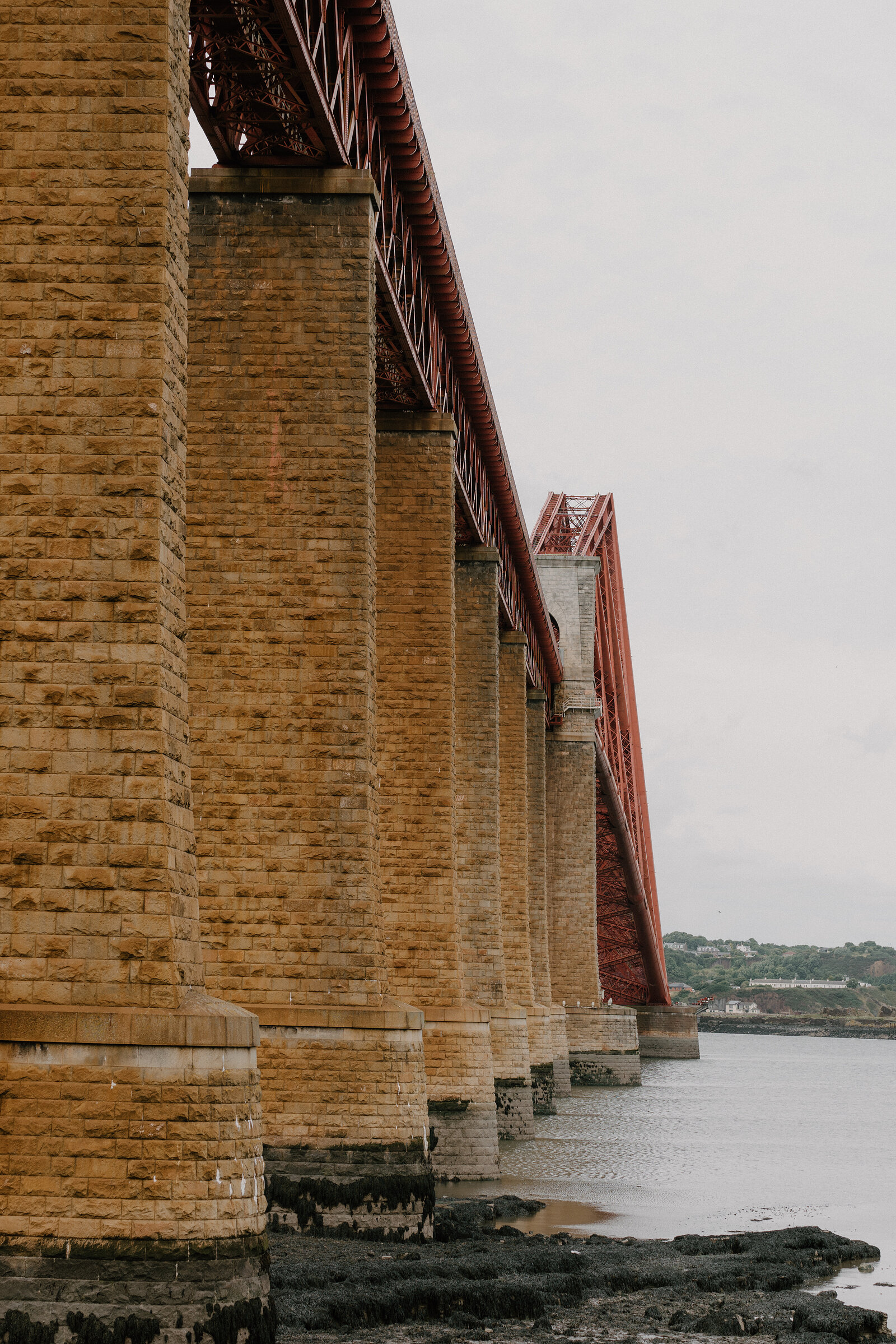The Forth Bridge's stone piers receding along the shoreline beneath its red cantilevered spans
