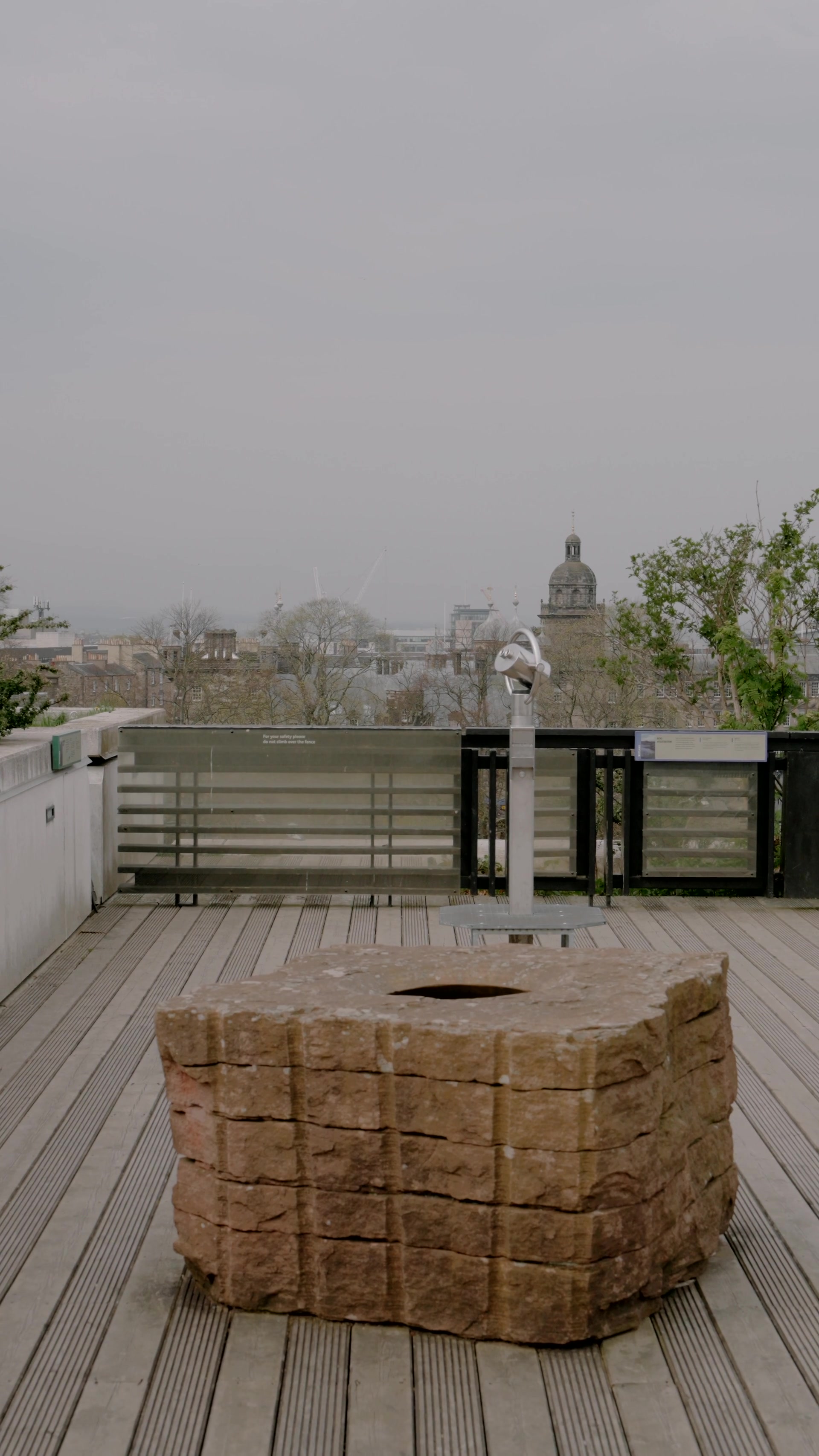 a rooftop terrace with a coin-operated telescope and a carved stone block overlooks a hazy city skyline
