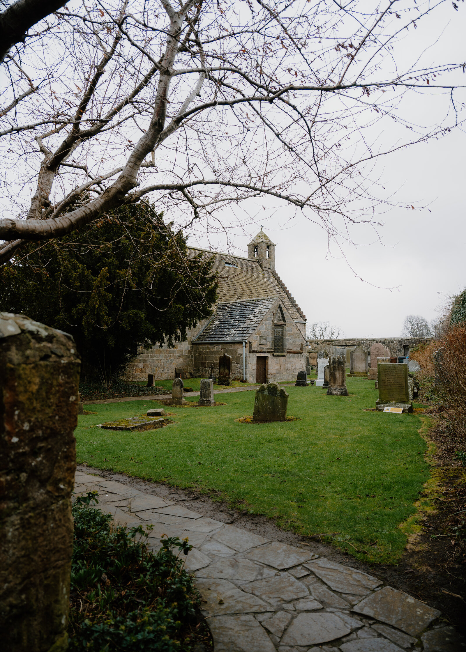 A small stone chapel with a bellcote stands behind a scatter of gravestones, a gravel path leading through the churchyard beneath bare branches.