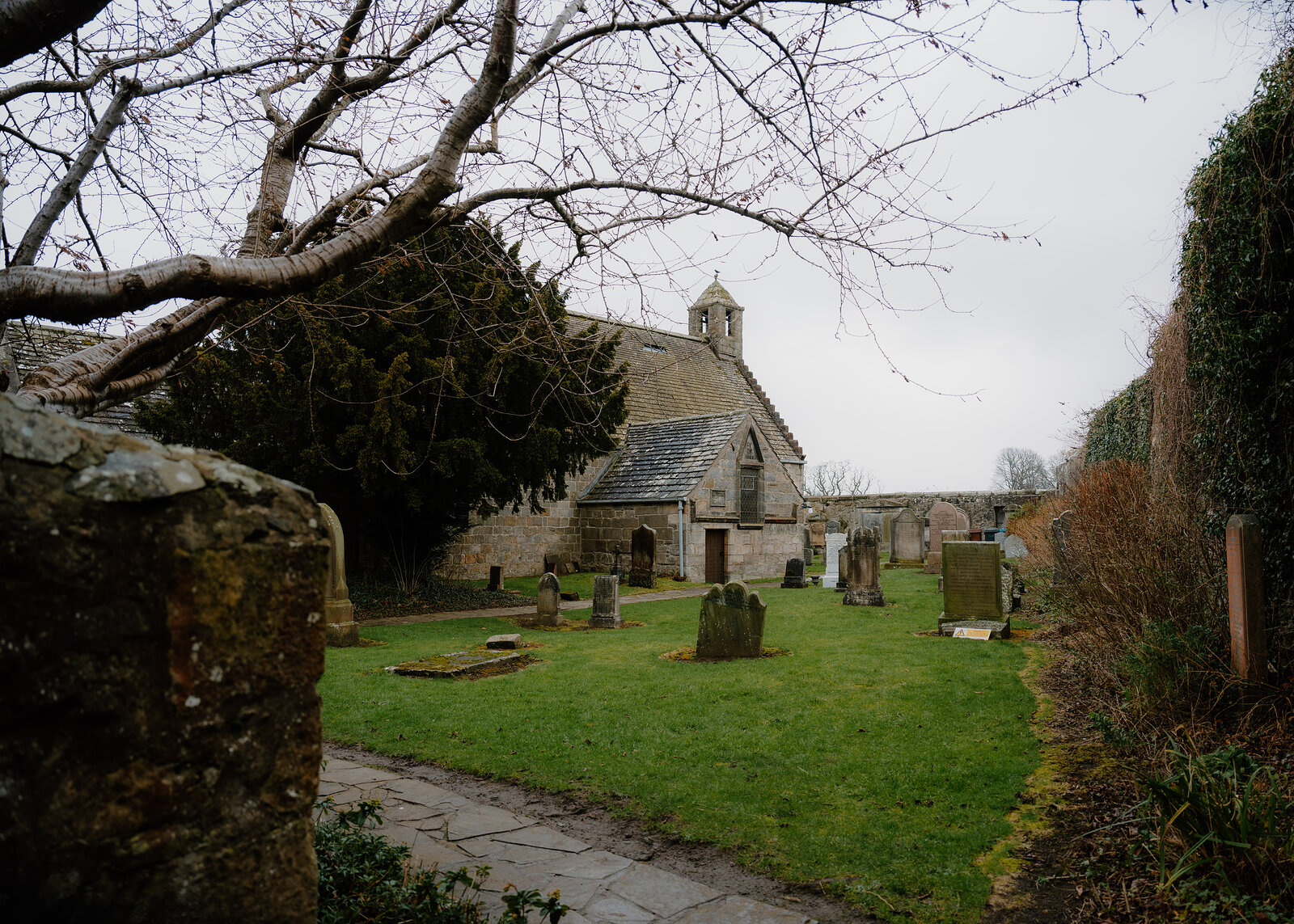 A small stone chapel with a bellcote stands beyond a low wall and gate, bare branches framing the view across the churchyard.