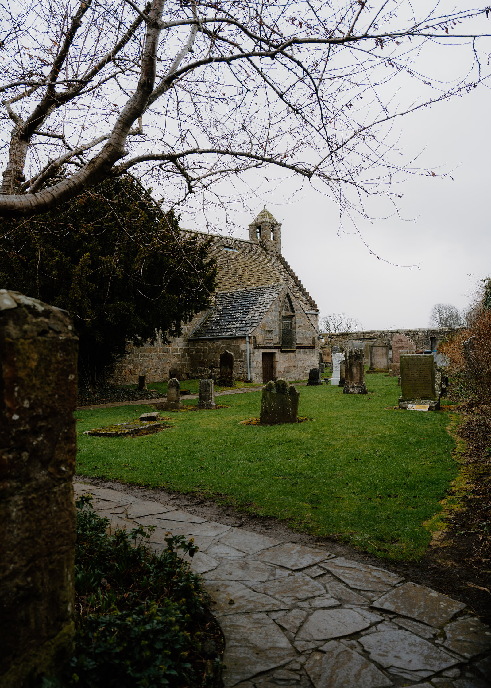 A small stone chapel with a bellcote rises behind gravestones, a paved path leading through the churchyard beneath bare branches.