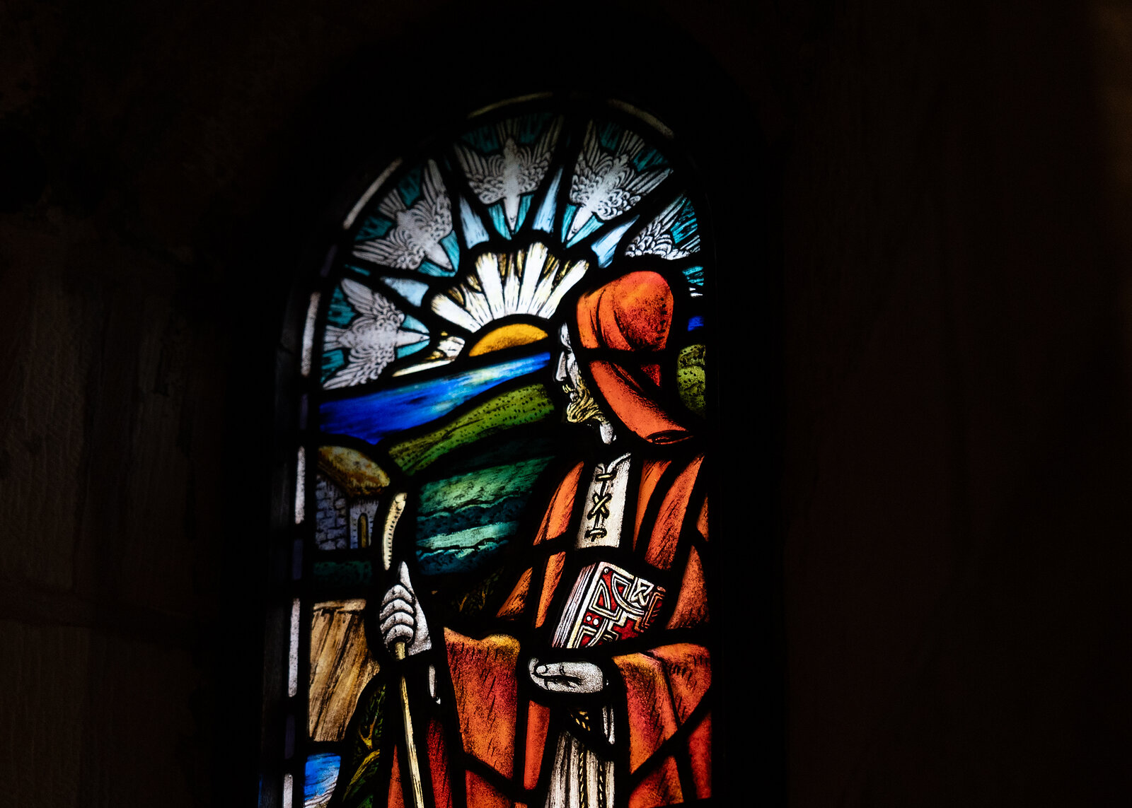 A stained-glass window shows a robed pilgrim with a staff standing before a sunrise over hills and sea, set into a dark stone archway.