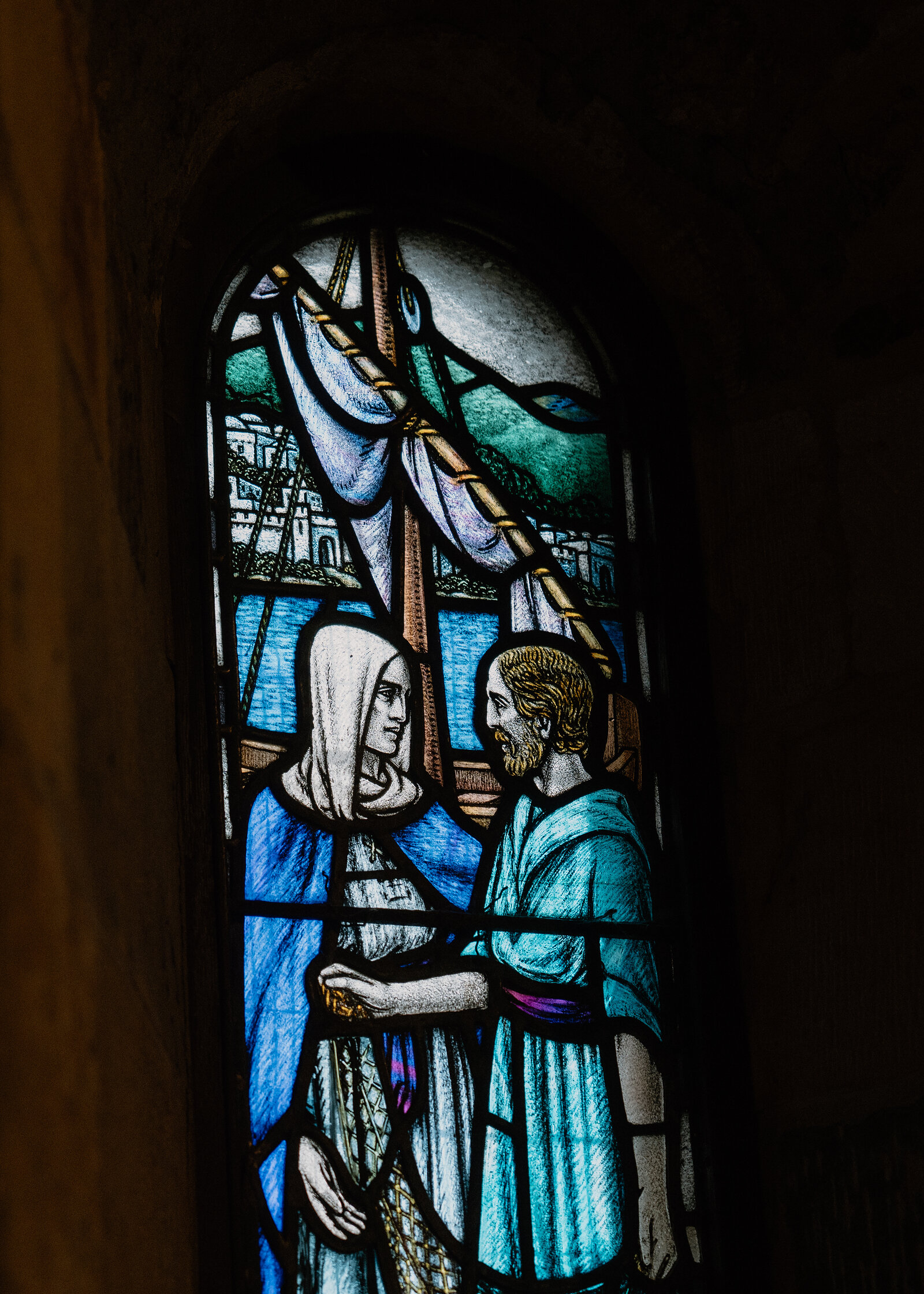 A stained-glass window shows two robed figures in conversation before a sailing boat and hills, set into a dark stone archway.