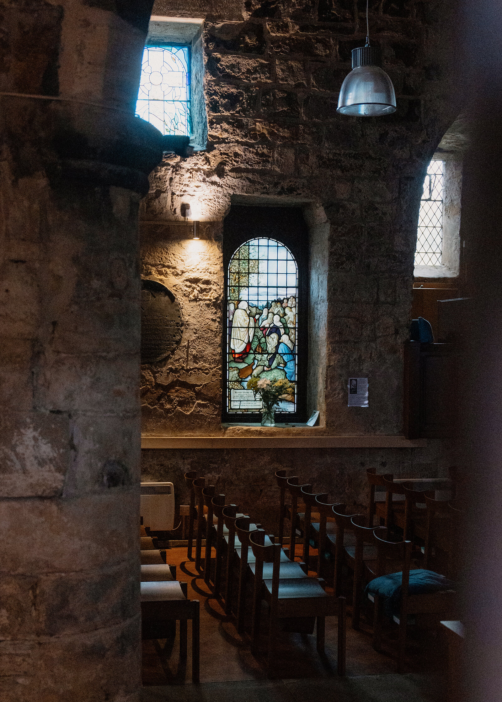 Rows of wooden chairs face a stained-glass window set into a thick stone wall inside a dimly lit chapel, a hanging light fitting above.