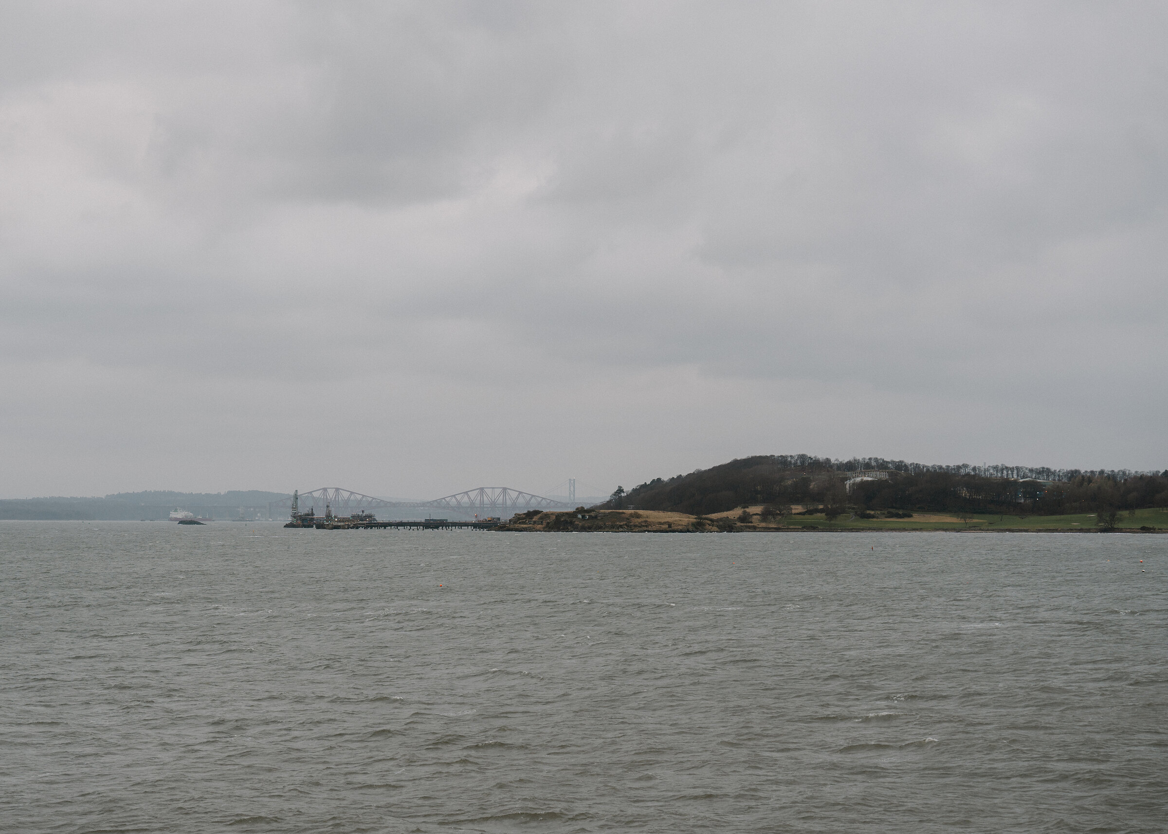A distant road bridge crosses the water beside a wooded island, calm sea stretching under a grey sky.