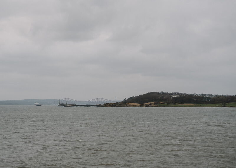 A distant road bridge crosses the water beside a wooded island, calm sea stretching under a grey sky.