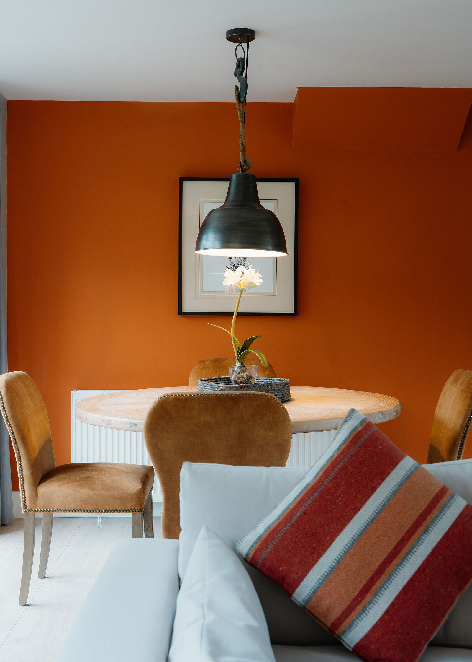 A black pendant lamp hangs over a white flower on a round table, with velvet chairs and a striped cushion in the foreground.