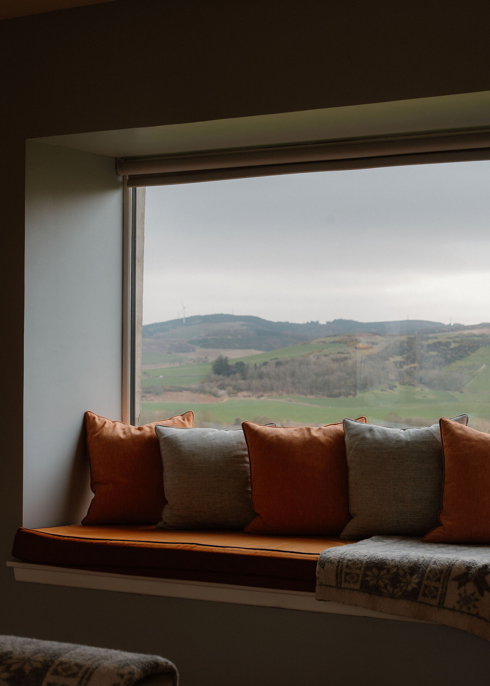 A window seat lined with orange and grey cushions and a knitted throw looks out over green hills and woodland.