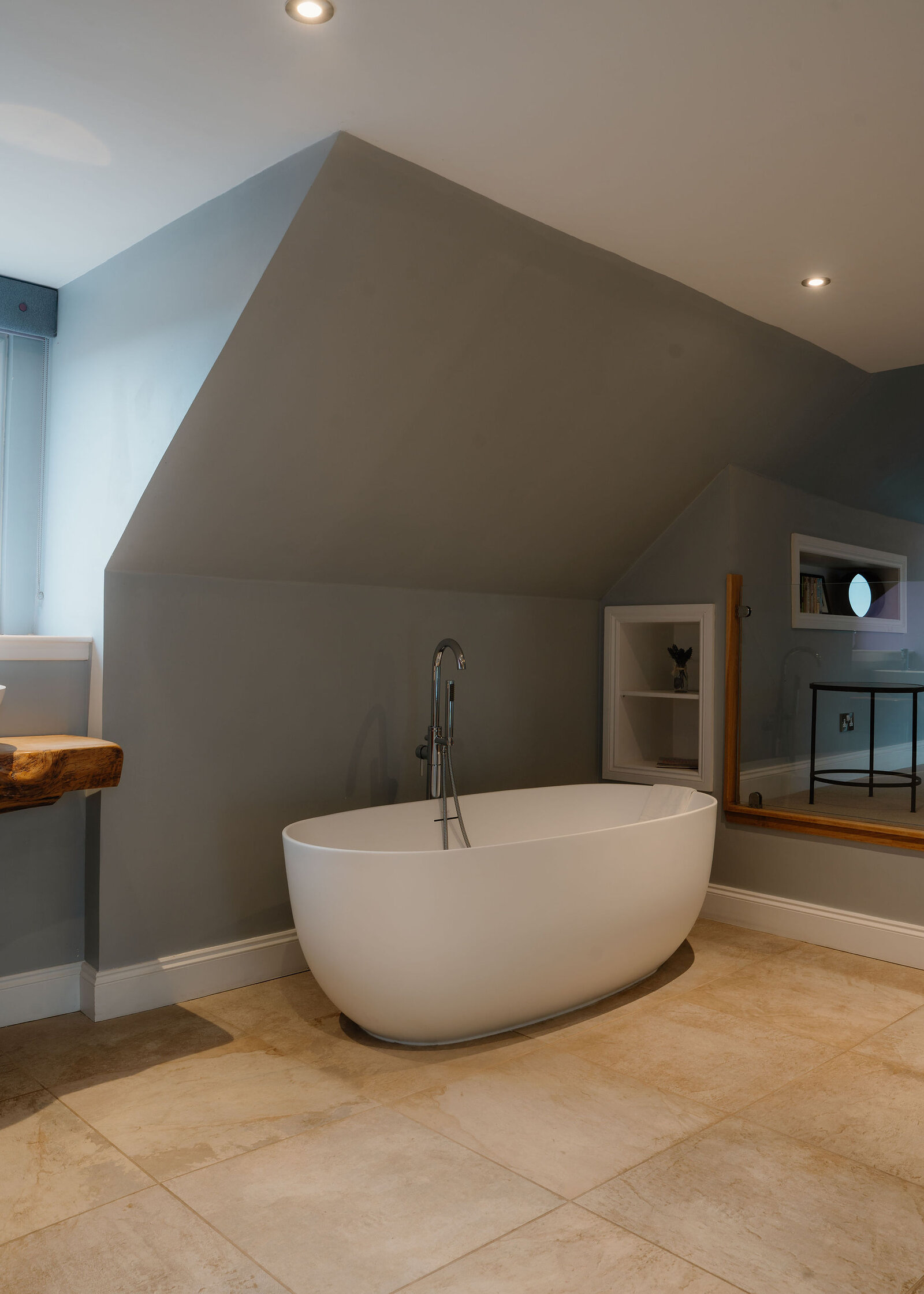 A freestanding bath stands below a sloped grey ceiling, a mirror reflecting a bedroom beyond a recessed shelf holding a small vase.