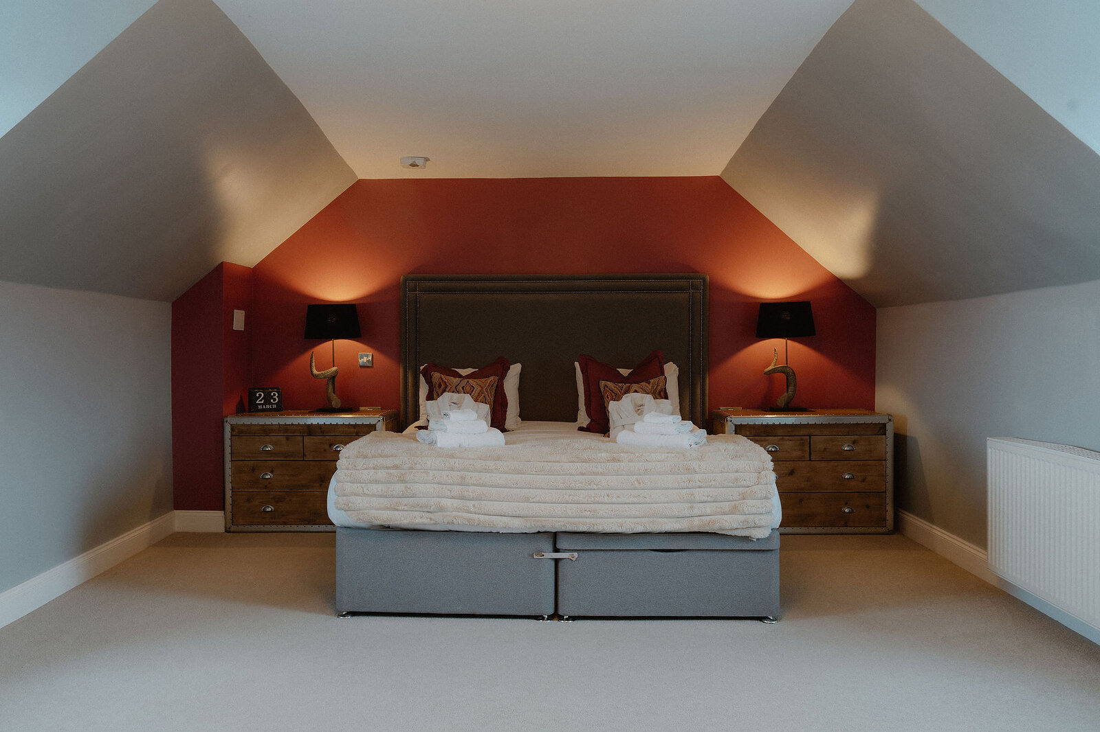 A double bed with a grey headboard stands against a terracotta end wall under a sloping ceiling, flanked by two horn lamps.
