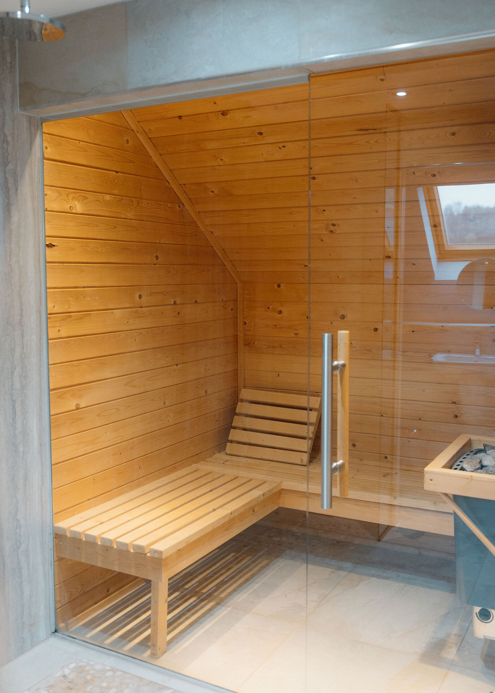 A pine-lined sauna with slatted benches and a heater of stones sits behind a glass door beside a rain shower.