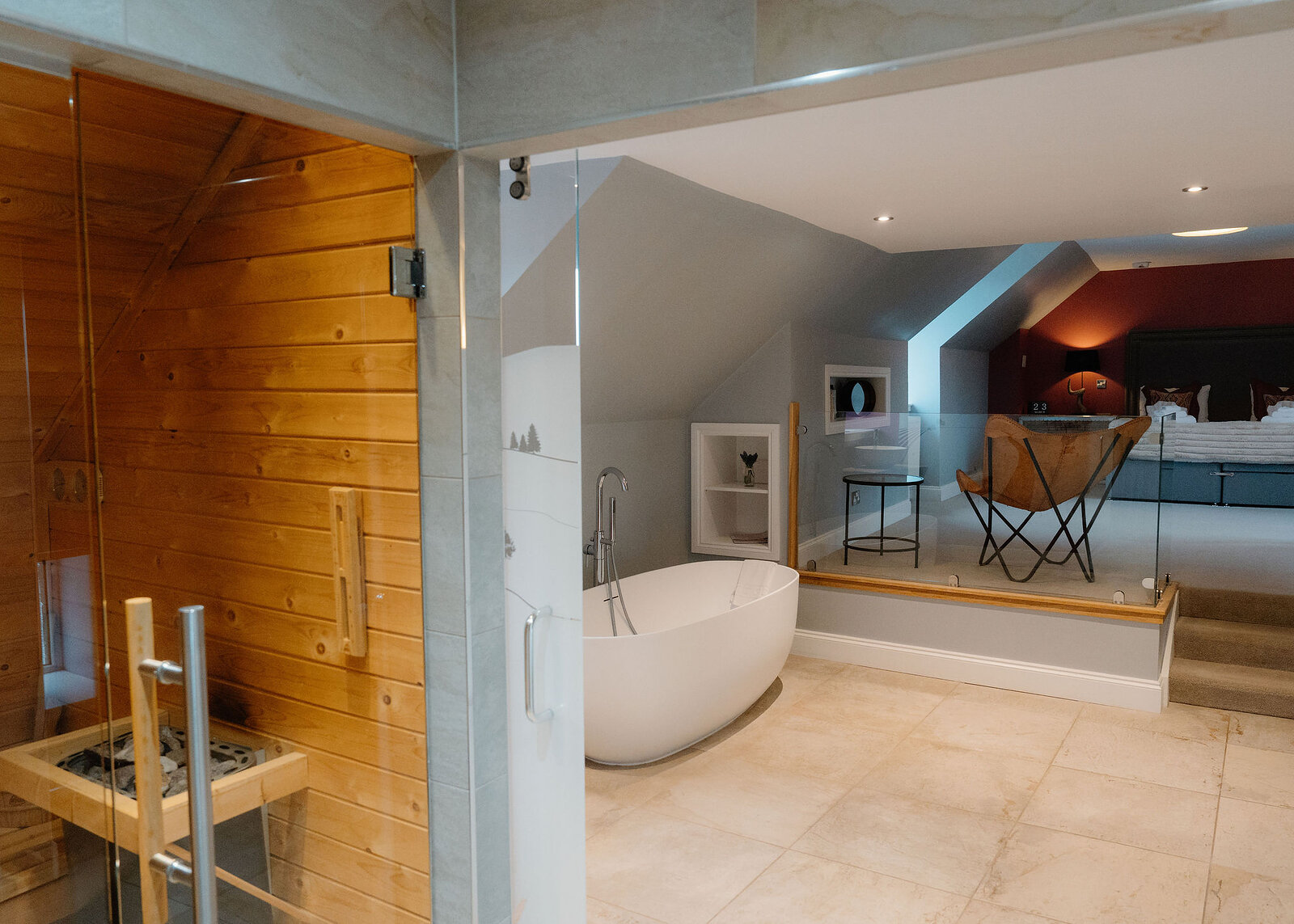A freestanding white bath stands between the sauna and a raised bedroom behind a glass balustrade with a leather butterfly chair.