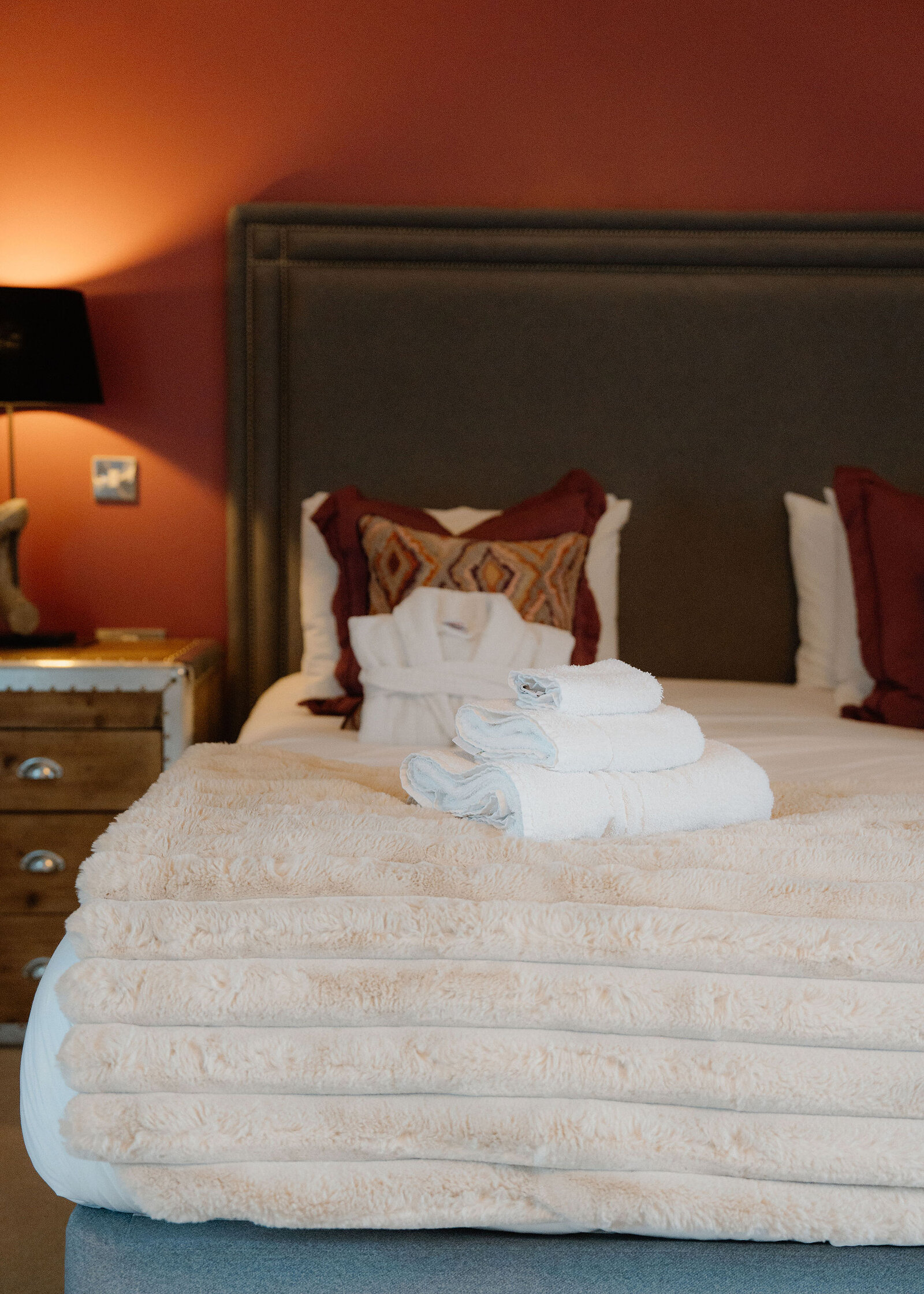 Folded white towels sit on a cream faux fur throw at the foot of a bed, with a bathrobe on the pillows behind.