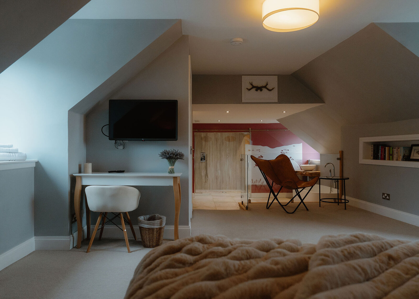 A white desk and chair sit beneath a mounted television, bookshelves to the right and a leather butterfly chair visible through the archway.