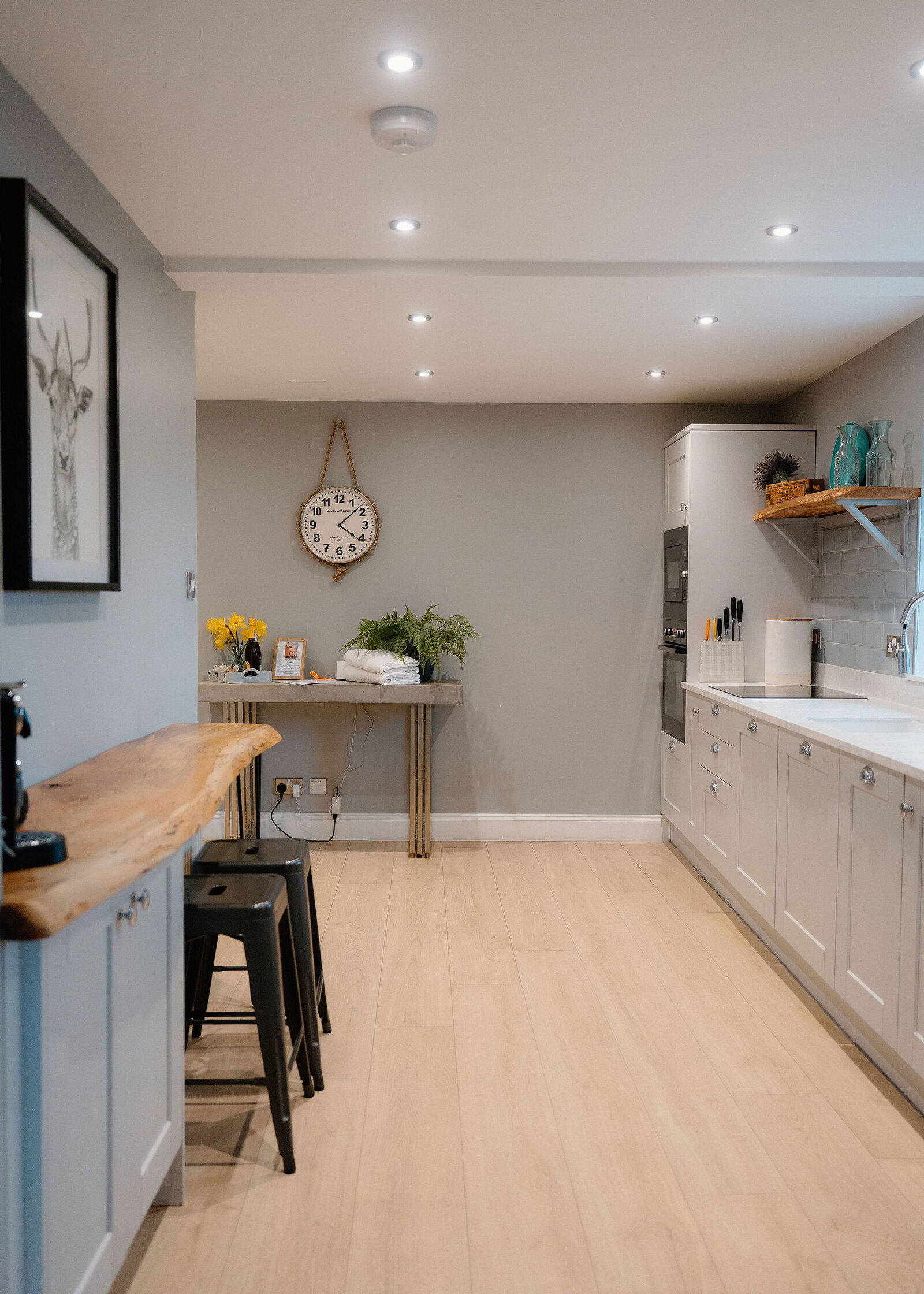 A round wall clock hangs above a narrow console table with flowers, black bar stools lining a breakfast bar beside a white fitted kitchen.