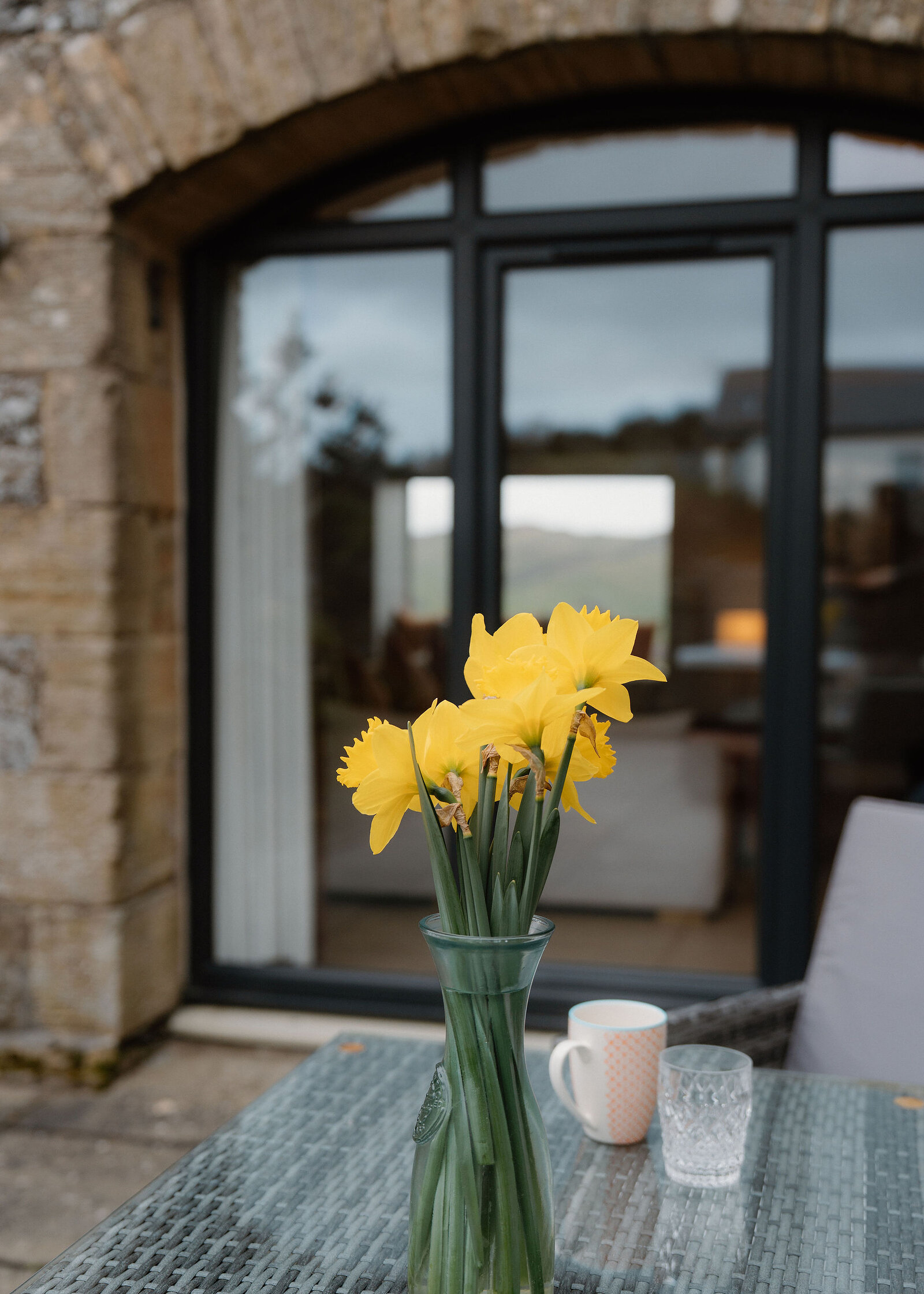 A vase of daffodils stands on an outdoor table before arched glazed doors, a mug and glass beside it reflecting a lit lamp inside.