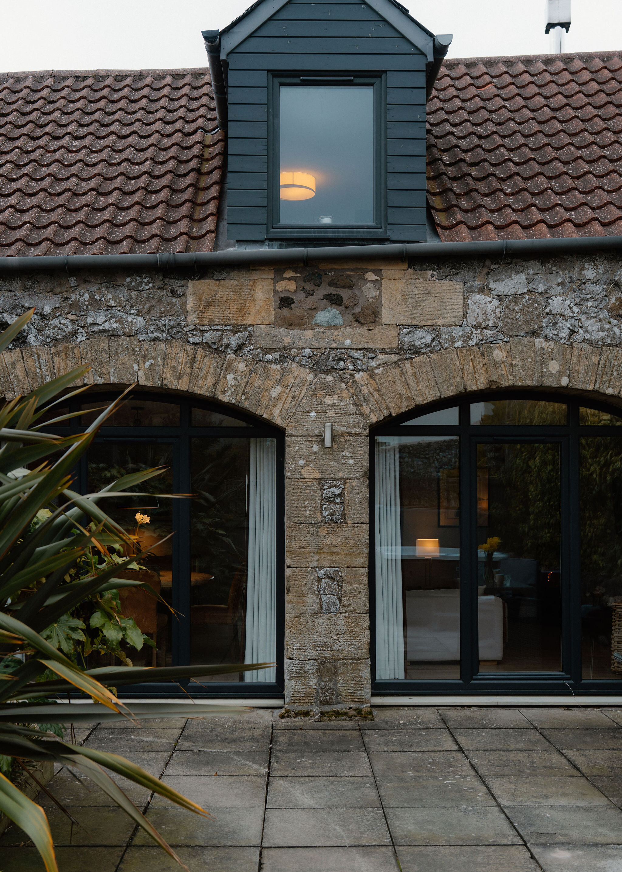 A stone cottage with a dark timber dormer and clay pantile roof stands above two arched glazed doors and a paved patio.