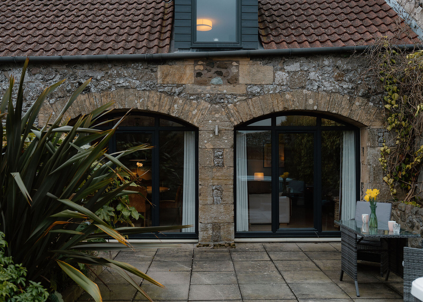 Two arched glazed doors in a stone wall open onto a paved patio, a rattan table with daffodils and mugs to the right.