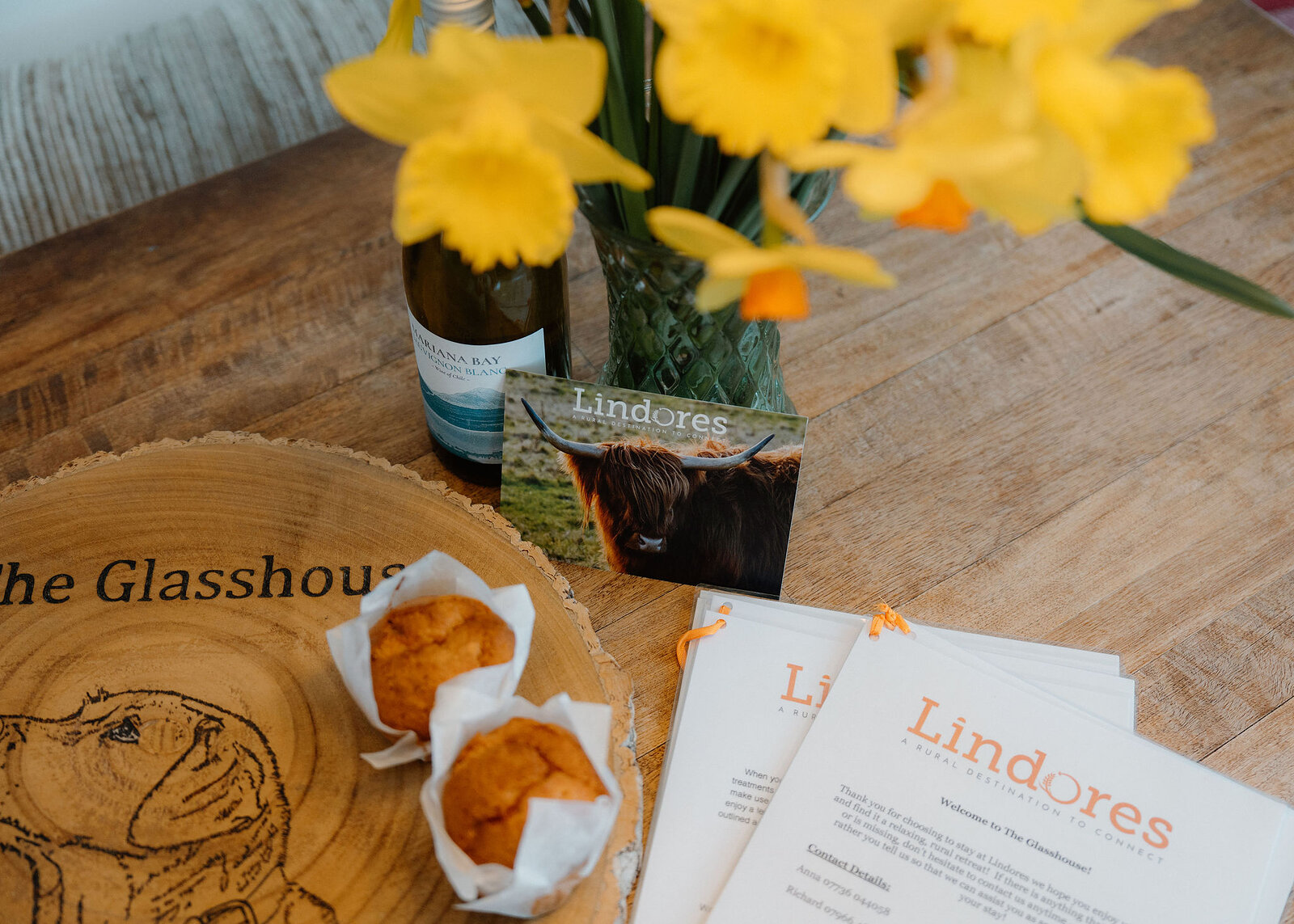 Two muffins sit on a log-slice board burned with The Glasshouse, seen from above beside Lindores welcome sheets, a Highland cow card and daffodils.
