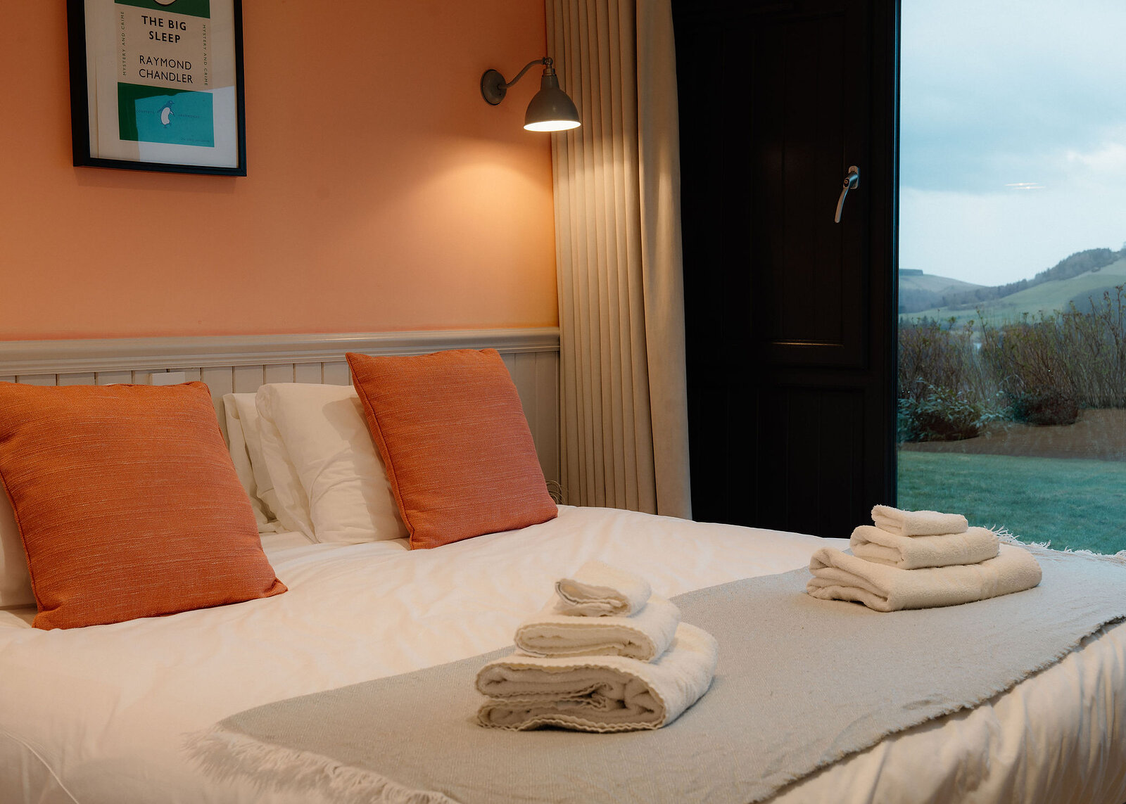 Folded towels sit on a bed with orange cushions against a peach wall, beside a glass door onto the garden and hills.