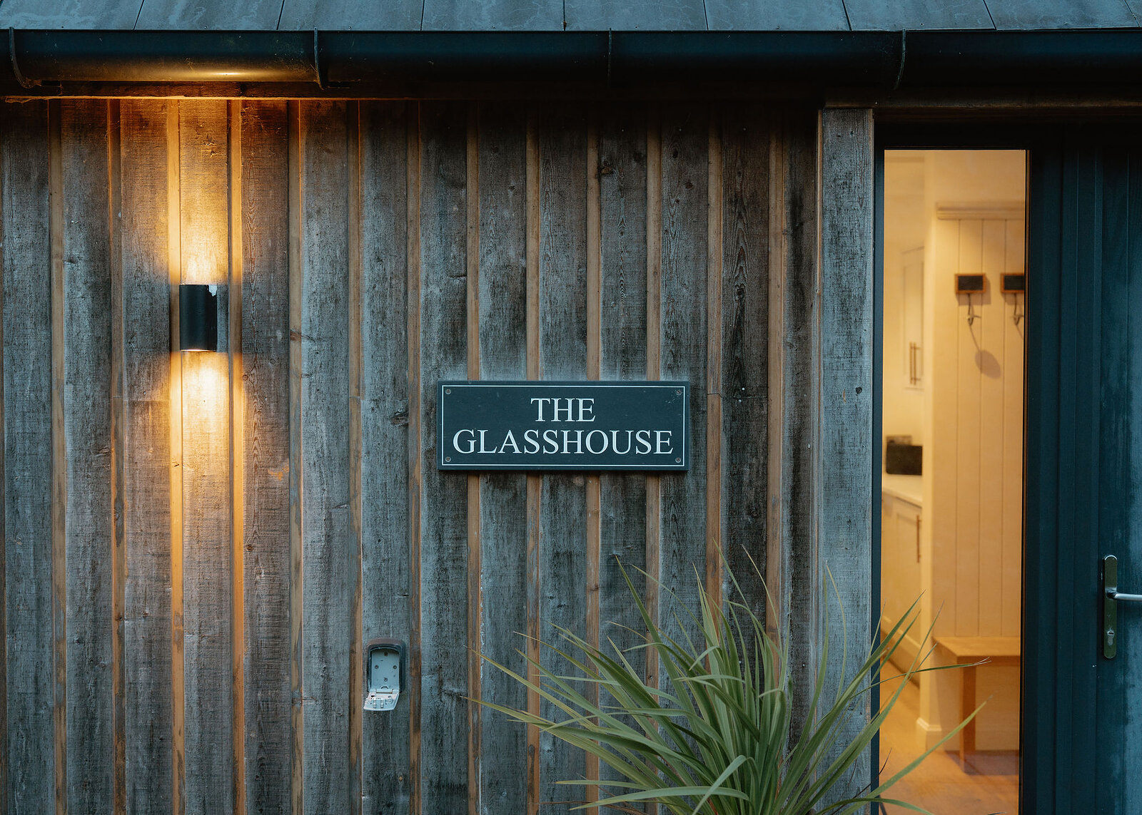 A slate sign reading The Glasshouse hangs on weathered timber cladding beside a lit wall light and an open door.
