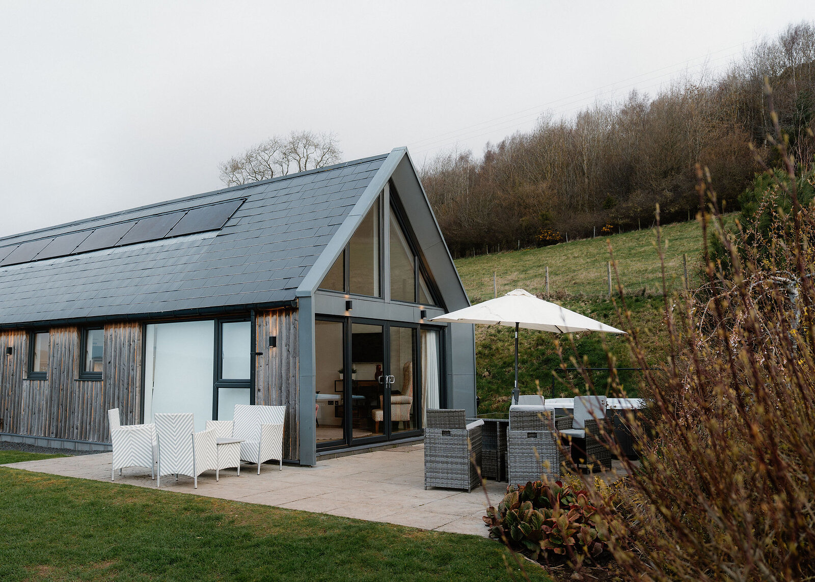 A timber house with a slate roof, solar panels and a glazed gable end opens onto a paved terrace with chairs and a parasol.