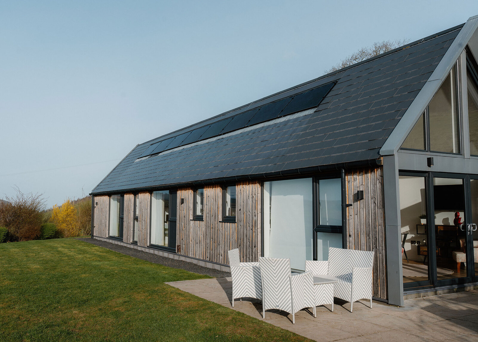 The long timber-clad side of the house runs under a slate roof with solar panels, white chairs on the terrace and a clear blue sky.