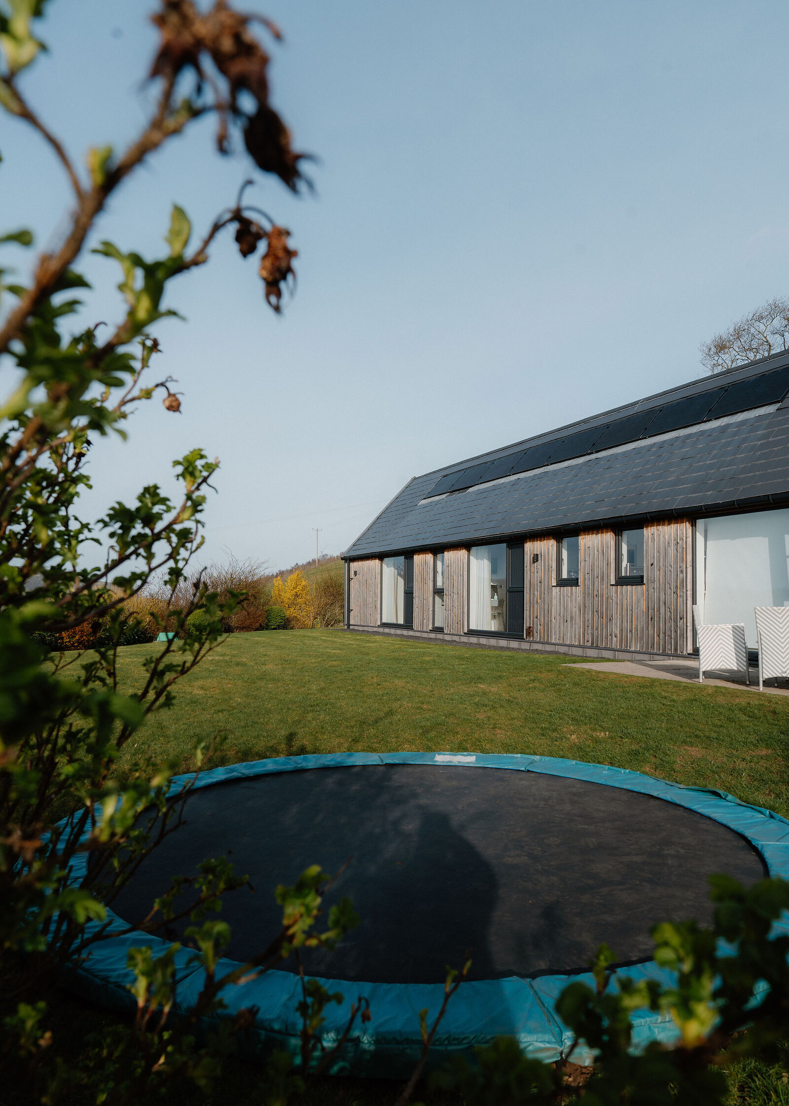A sunken trampoline sits on a lawn beside a long timber-clad house with solar panels, white chairs on its terrace.