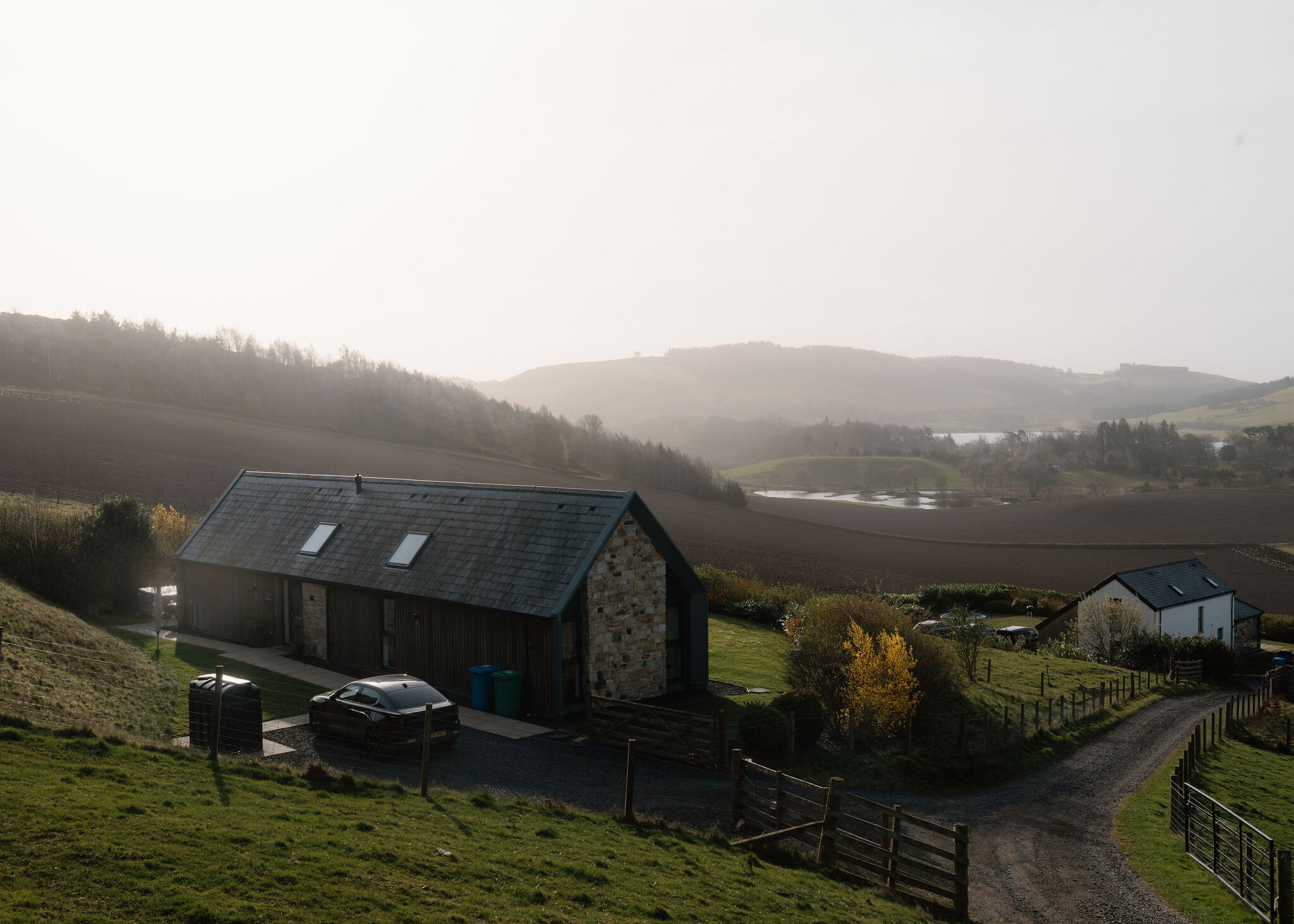 A stone and timber barn-style house sits in a valley of ploughed fields, mist settling over the hills and a small loch in the distance.