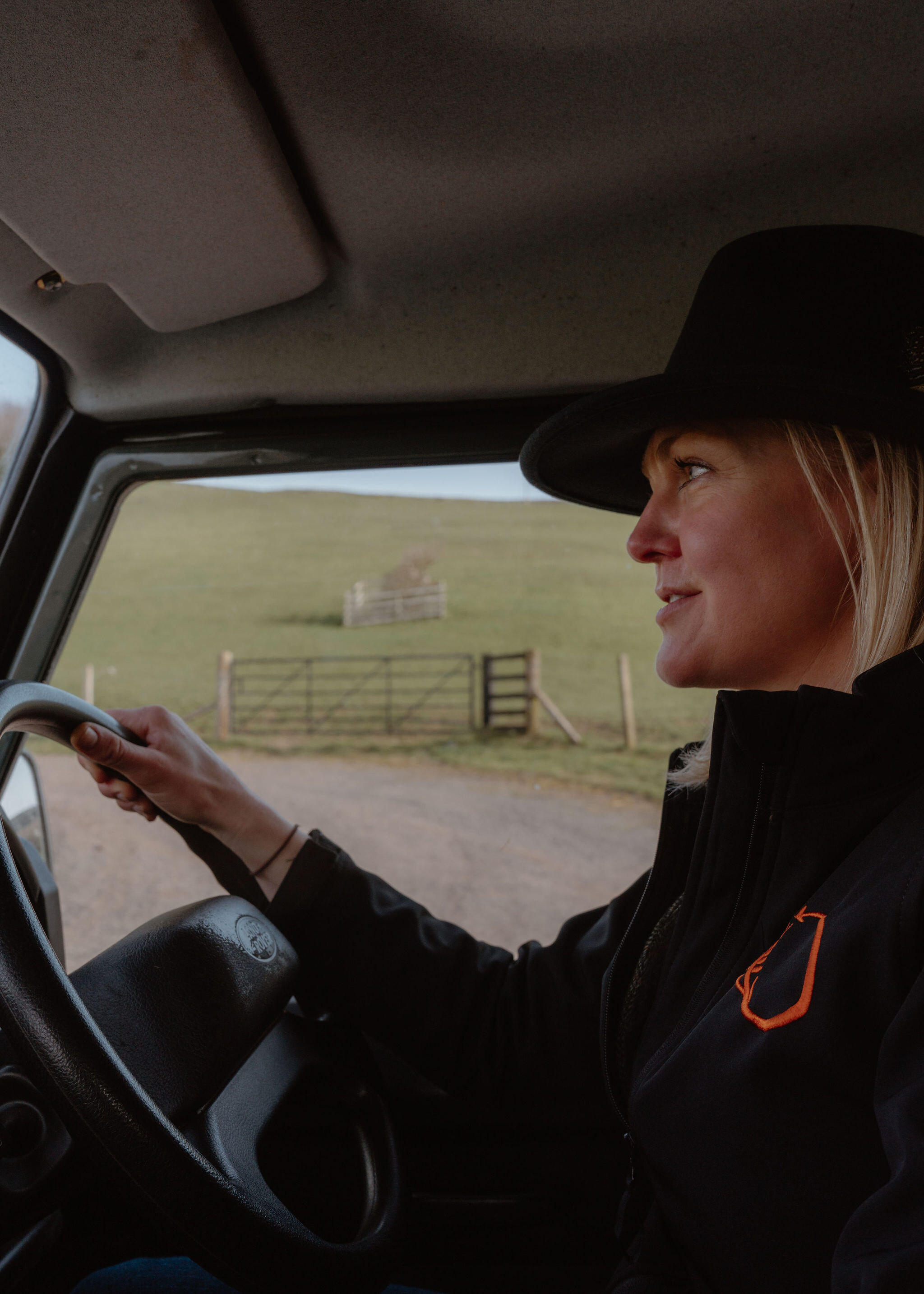 A woman in a wide-brimmed hat drives the Land Rover, one hand on the wheel, a field gate and wooden jump through the window.