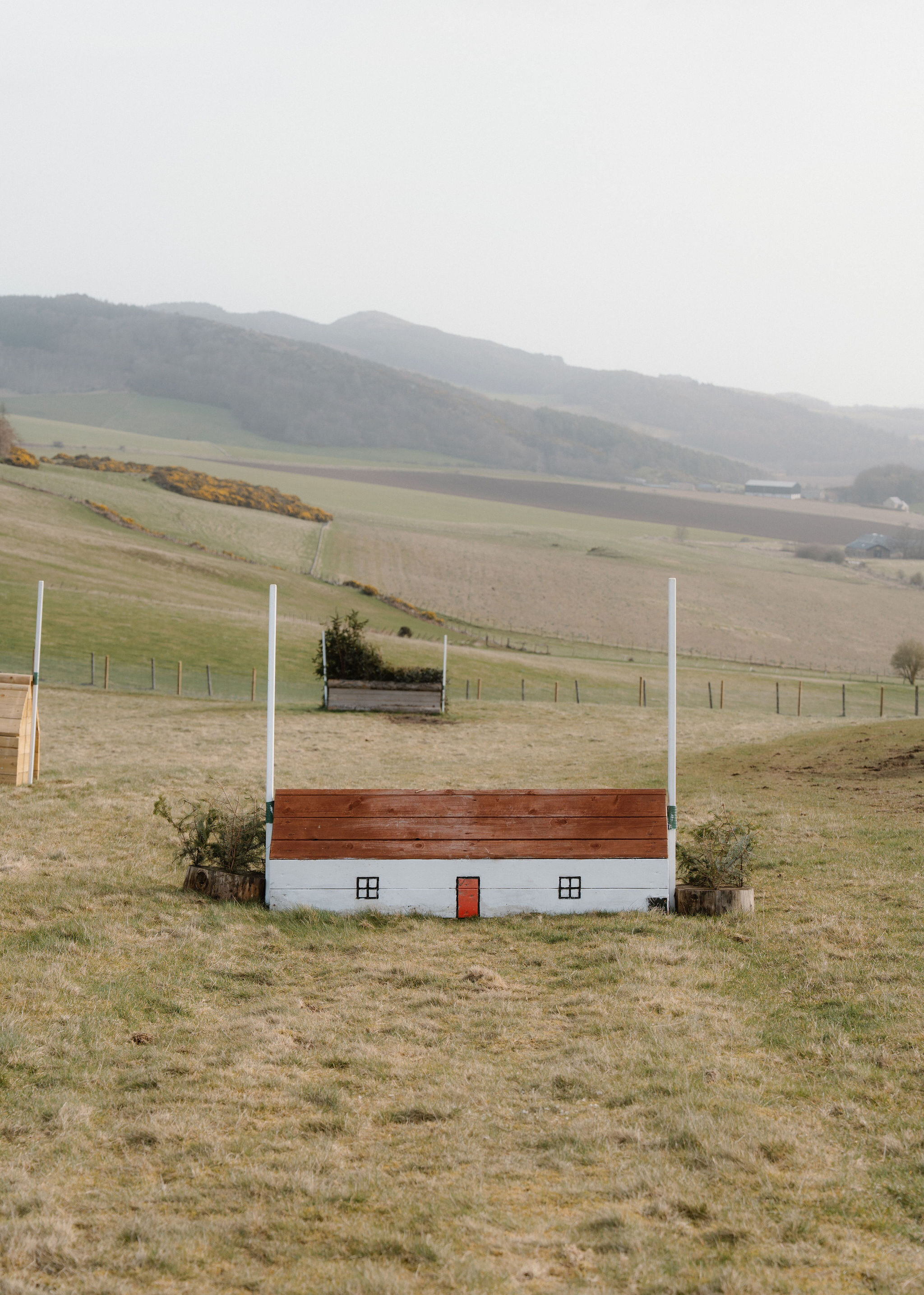 A cross-country jump built like a white cottage, with a red roof, red door and two windows, stands between white poles.
