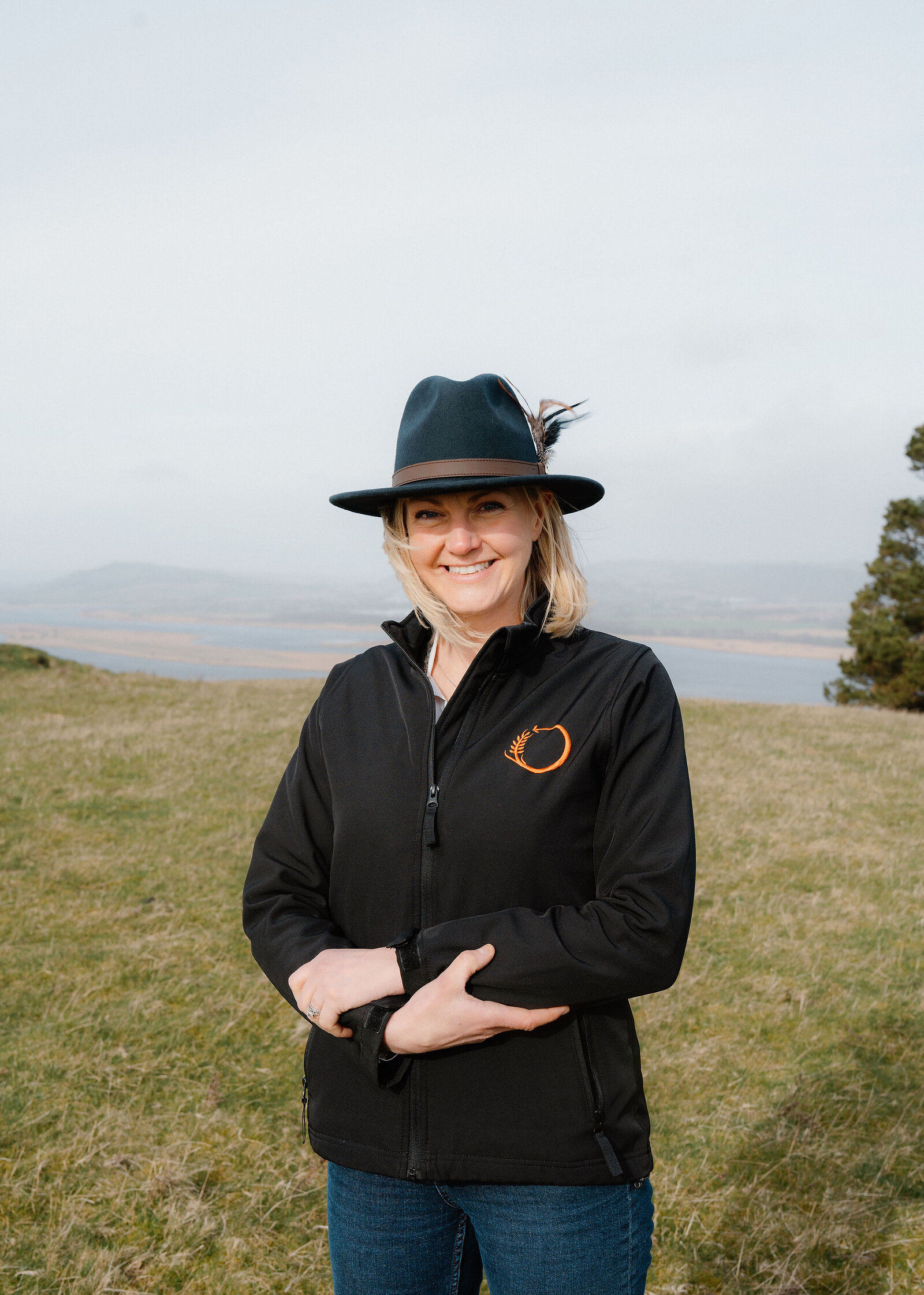 A smiling woman in a feathered wide-brimmed hat and black jacket faces the camera on a grassy hilltop above the misty River Tay.