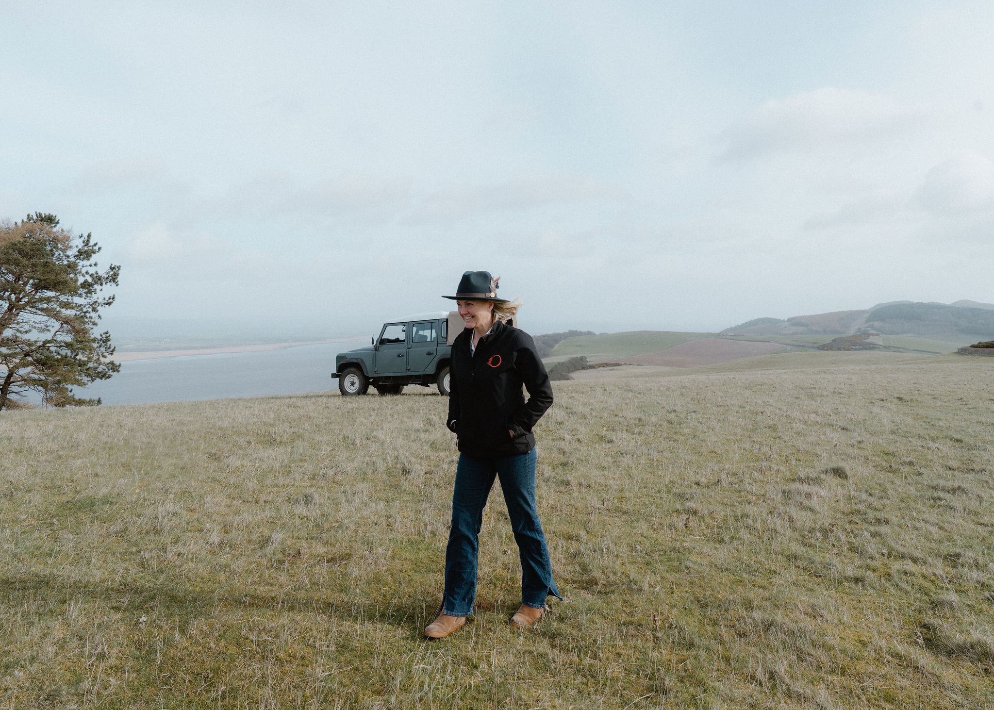 A woman in a wide-brimmed hat walks across the rough hilltop grass, smiling, with the parked Land Rover Defender and a Scots pine behind.