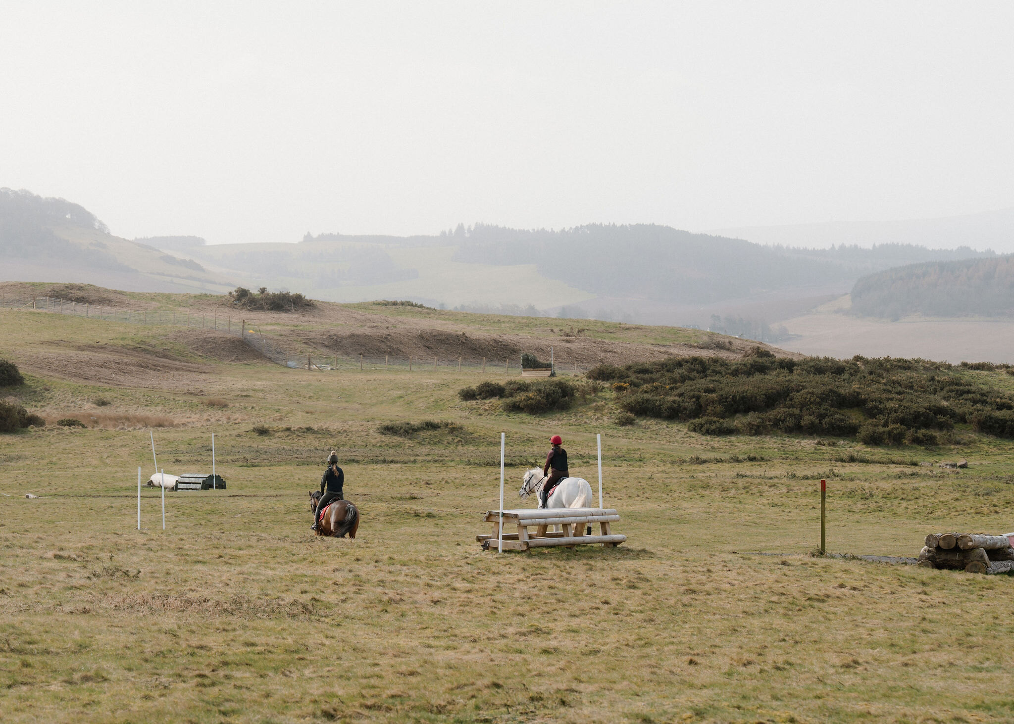 Two riders on a bay and a grey horse, seen from behind, approach a timber table jump across the rough cross-country course beneath misty hills.