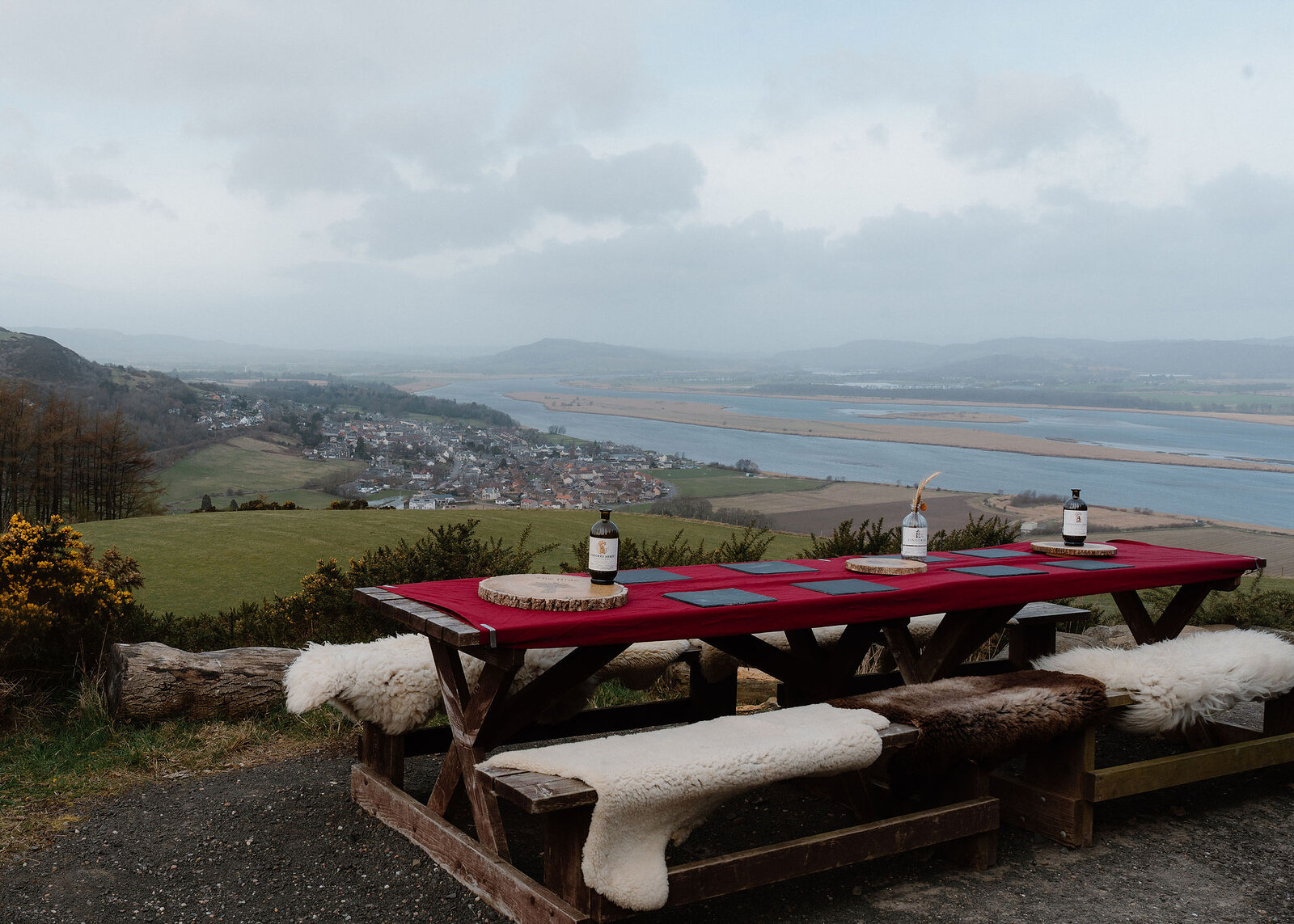 A long table under a red cloth, laid with slate mats, log slices and three bottles, its sheepskin benches above Newburgh.