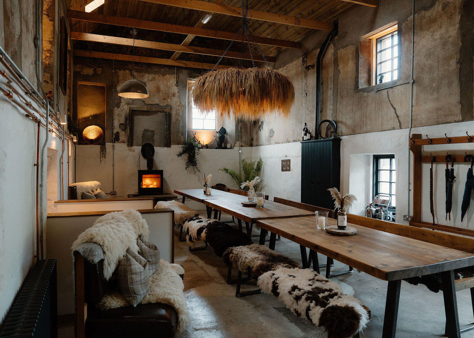 Long timber tables with sheepskin benches run towards a lit stove under a dried-grass chandelier, with umbrellas hanging on a coat rack at right.