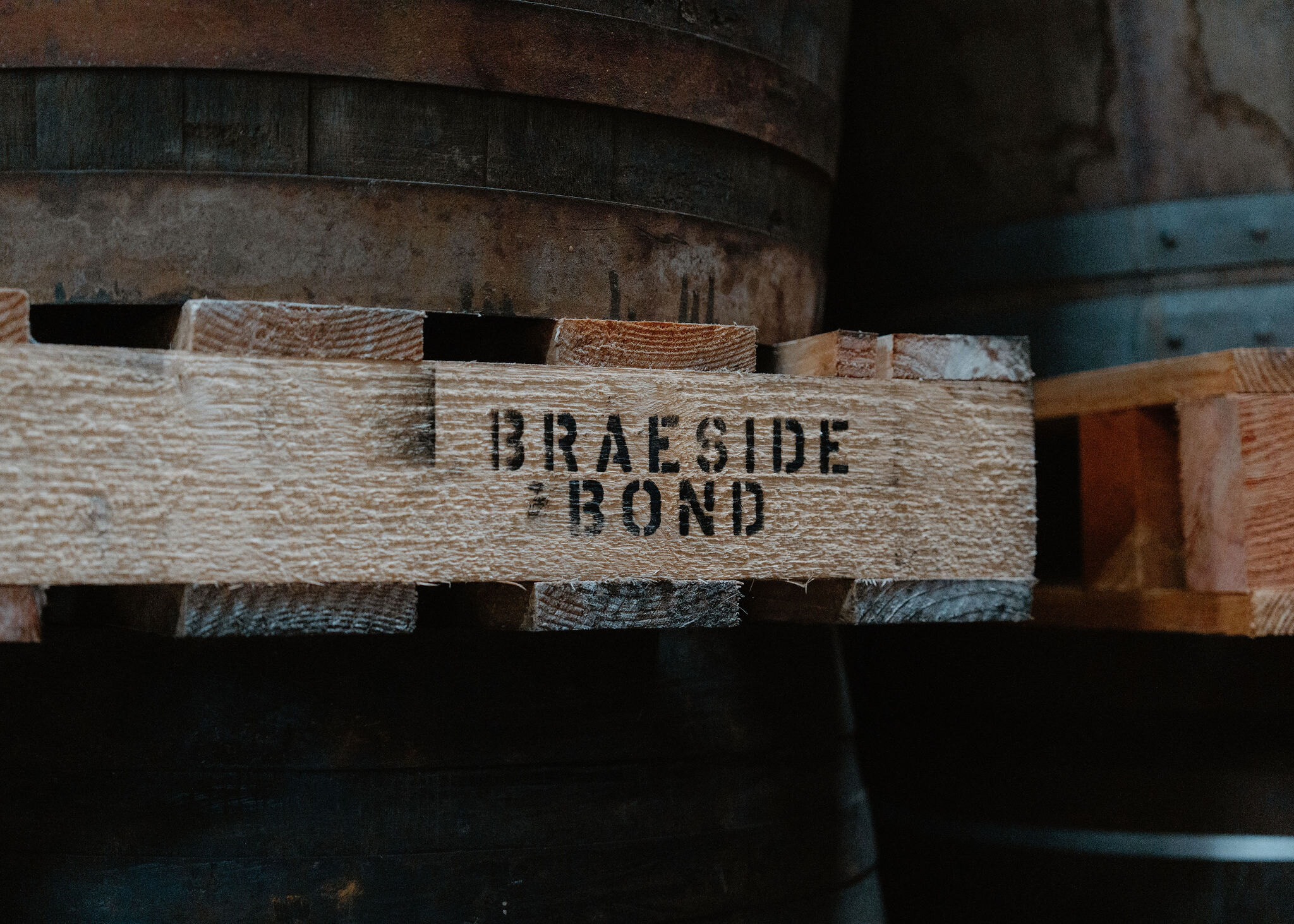 A close-up of the words Braeside Bond stencilled in black on the rough timber of a pallet beneath a whisky cask.