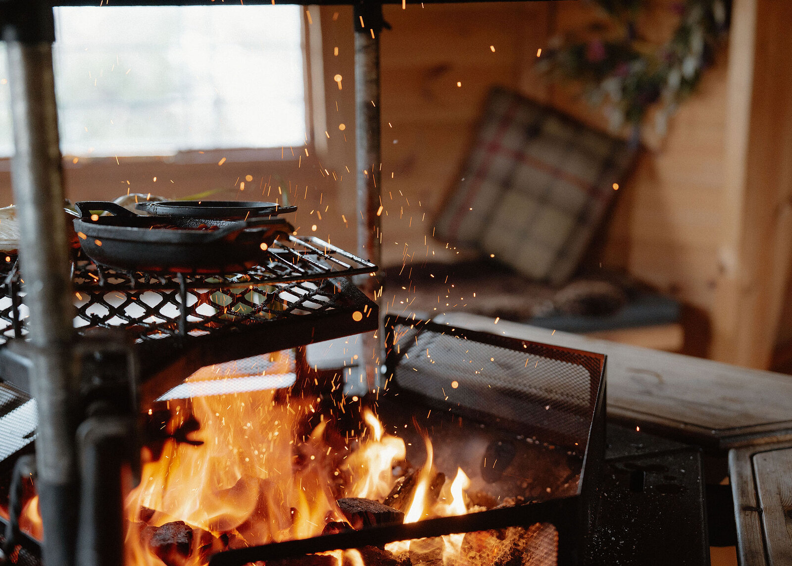 Sparks fly up from the open fire past two cast-iron pans on the grill rack, a window blurred in the hut behind.