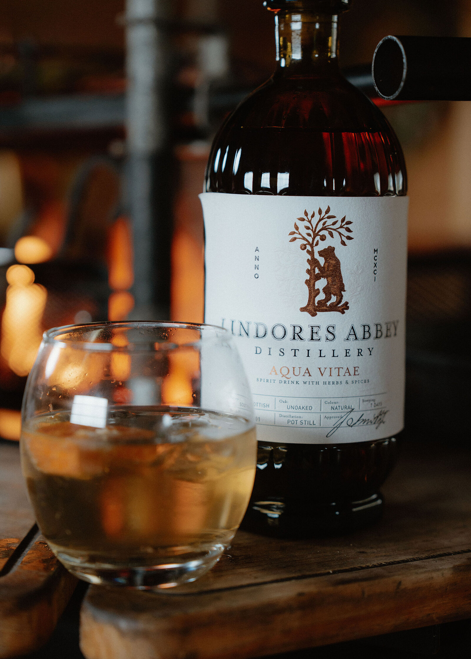 A Lindores Abbey Distillery Aqua Vitae bottle stands beside a tumbler of pale spirit and ice on a wooden ledge, the fire glowing behind.