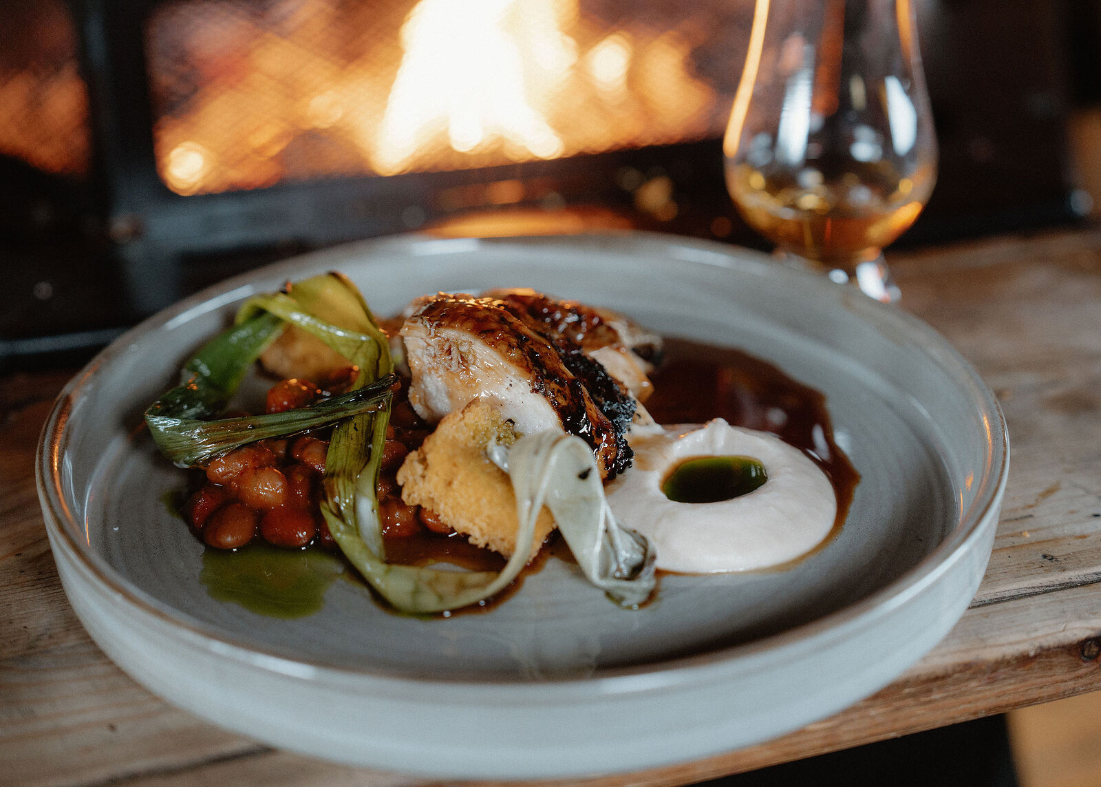 A low, close landscape view of the plate, the purée and green oil sharp and the fire and whisky glass blurred behind.