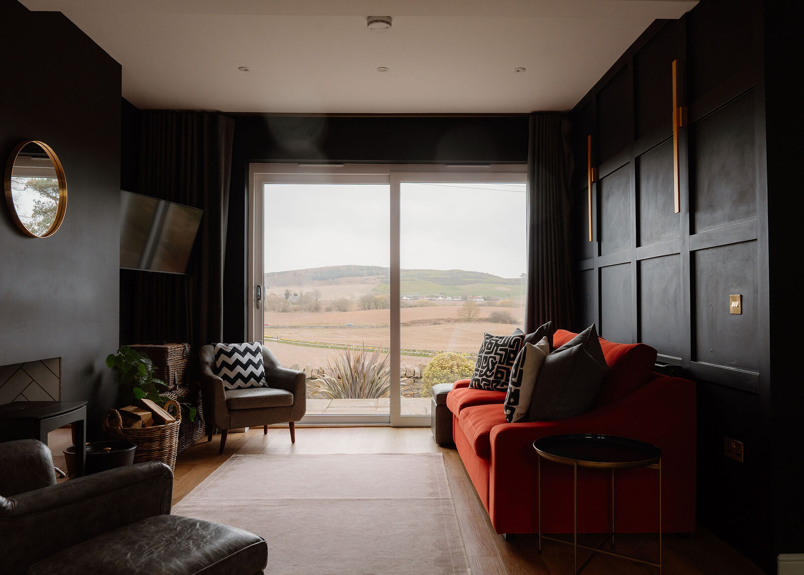 A dark living room with an orange sofa, grey armchair and leather chair looks through sliding glass doors to fields and a hill.
