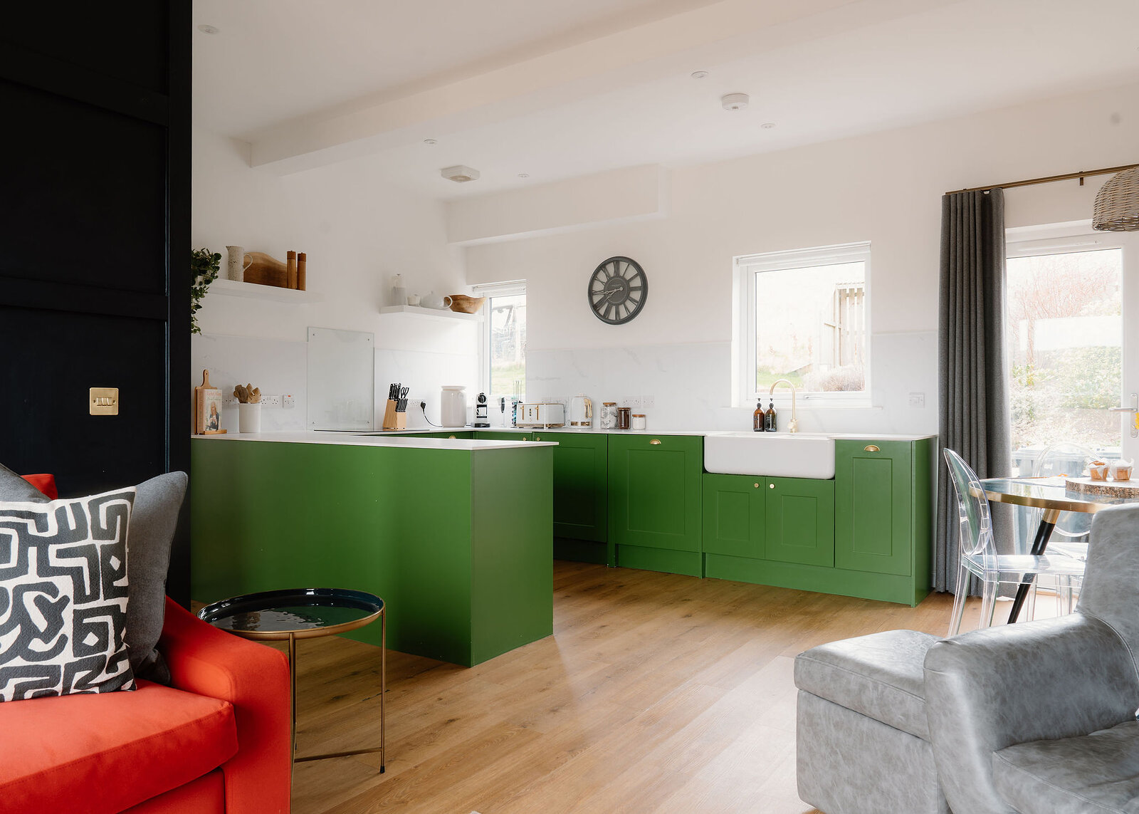An open-plan room pairs a green kitchen and Belfast sink with an orange sofa, grey leather chairs and a round glass dining table.